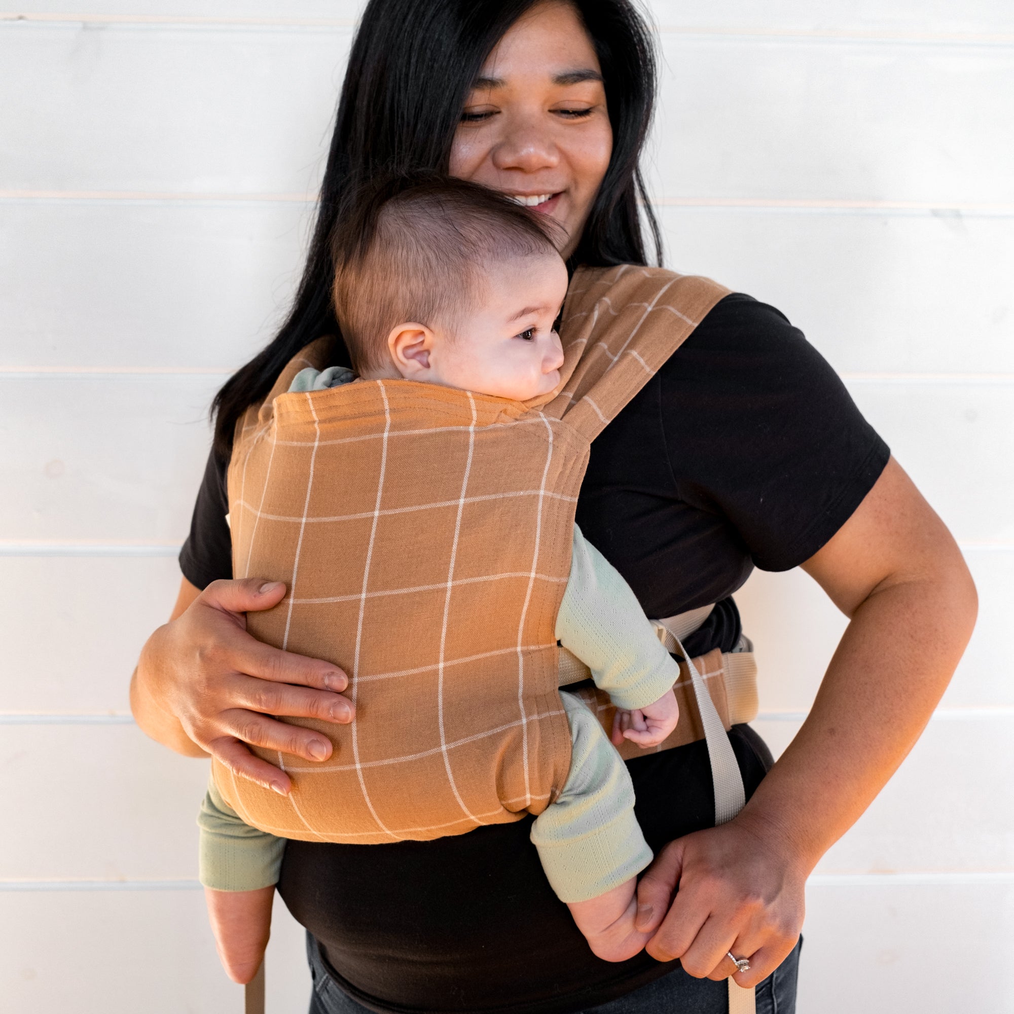 Woman holding a baby in a brown baby carrier with a white grid against a white background
