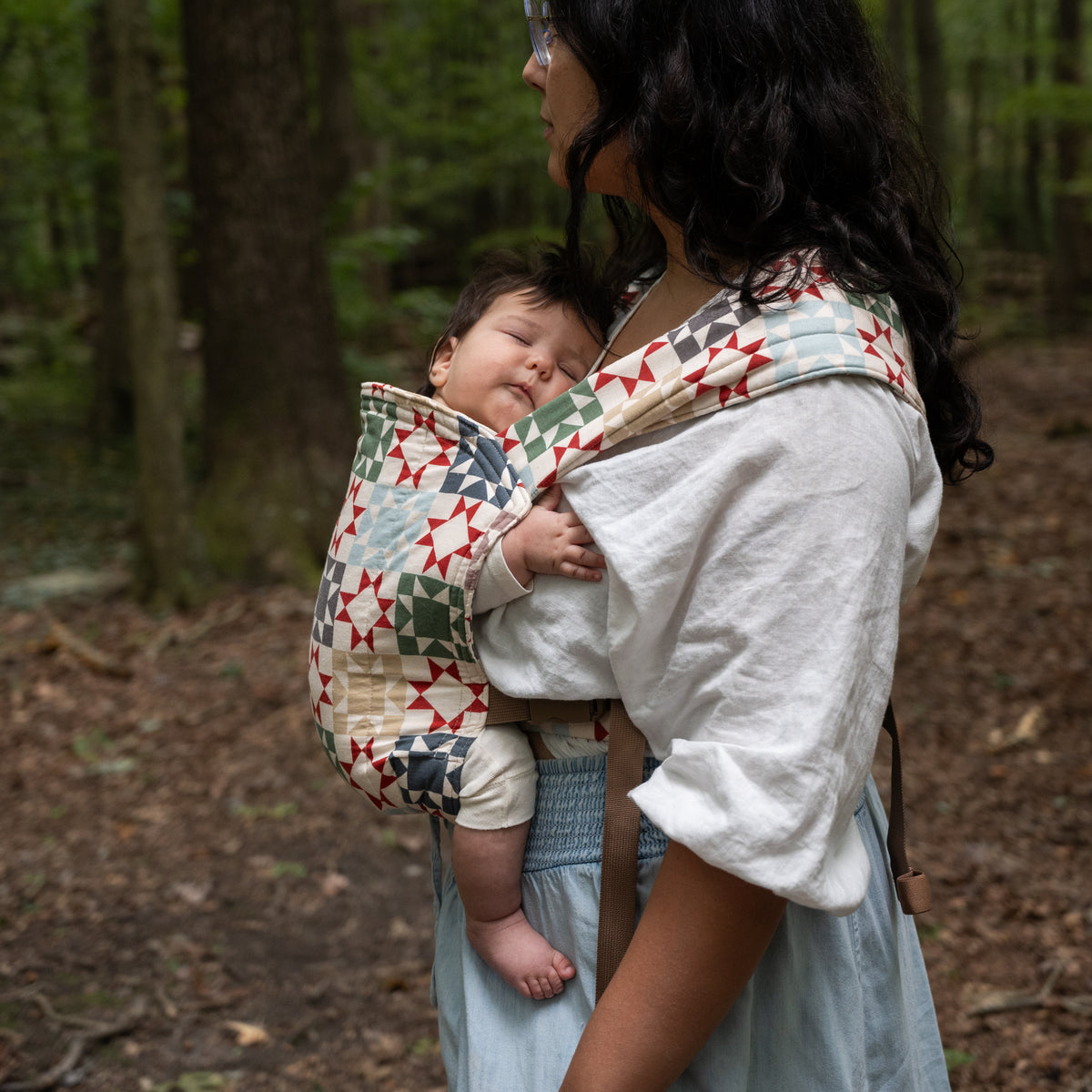A baby sleeps in the Heirloom Lark Baby Carrier, the quilted hood folded over the wearer's shoulder, showing star blocks in rust-red, sage, navy and tan on cream.