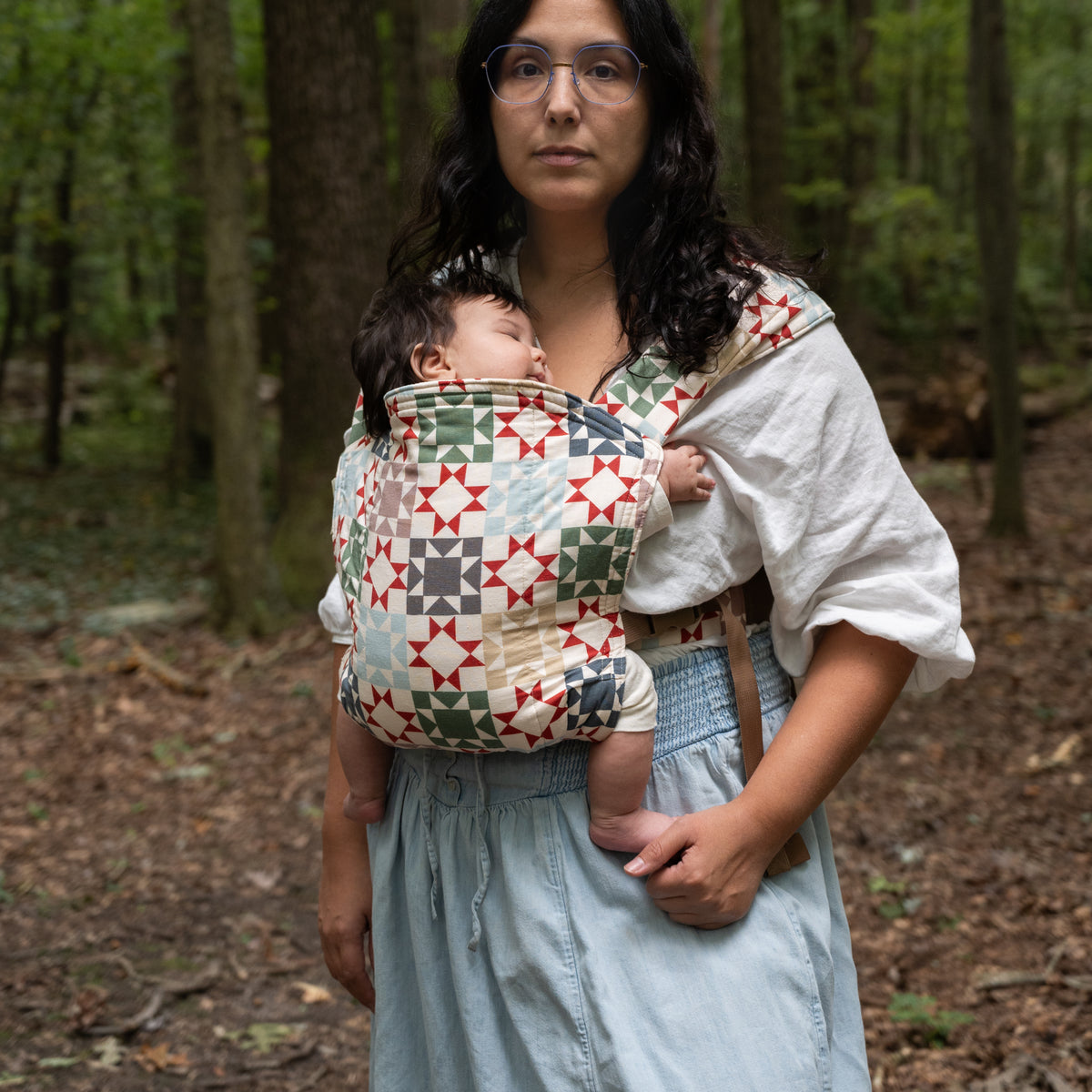 A wearer faces the camera with a sleeping baby in the Heirloom Lark Baby Carrier, the full panel of rust-red, sage, navy and tan quilt blocks on cream in view.