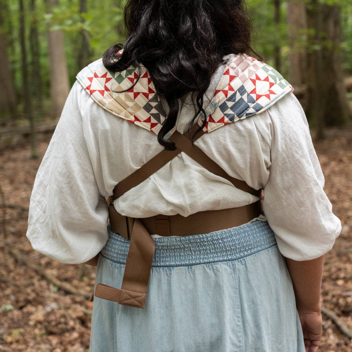 Back view of the Heirloom Lark Baby Carrier showing the crossed tan webbing straps, buckled waistband and quilted shoulder panels in the rust-red and navy star print.