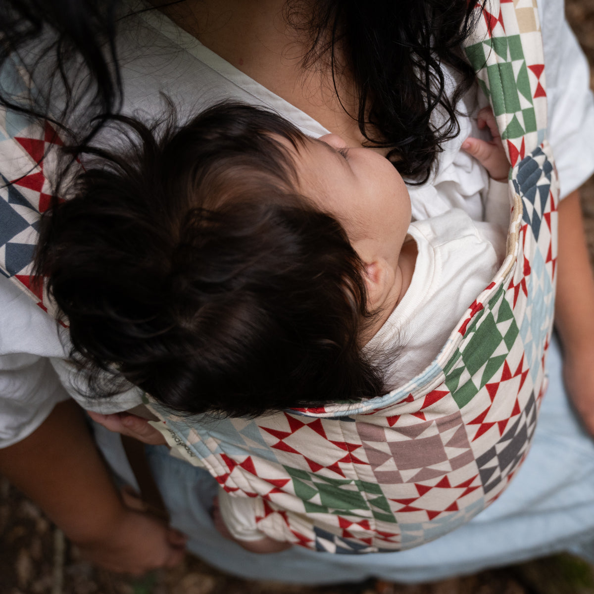 Close overhead view of a baby's head resting against the Heirloom panel, showing the quilt-block print of rust-red, sage, navy and tan triangles on cream.