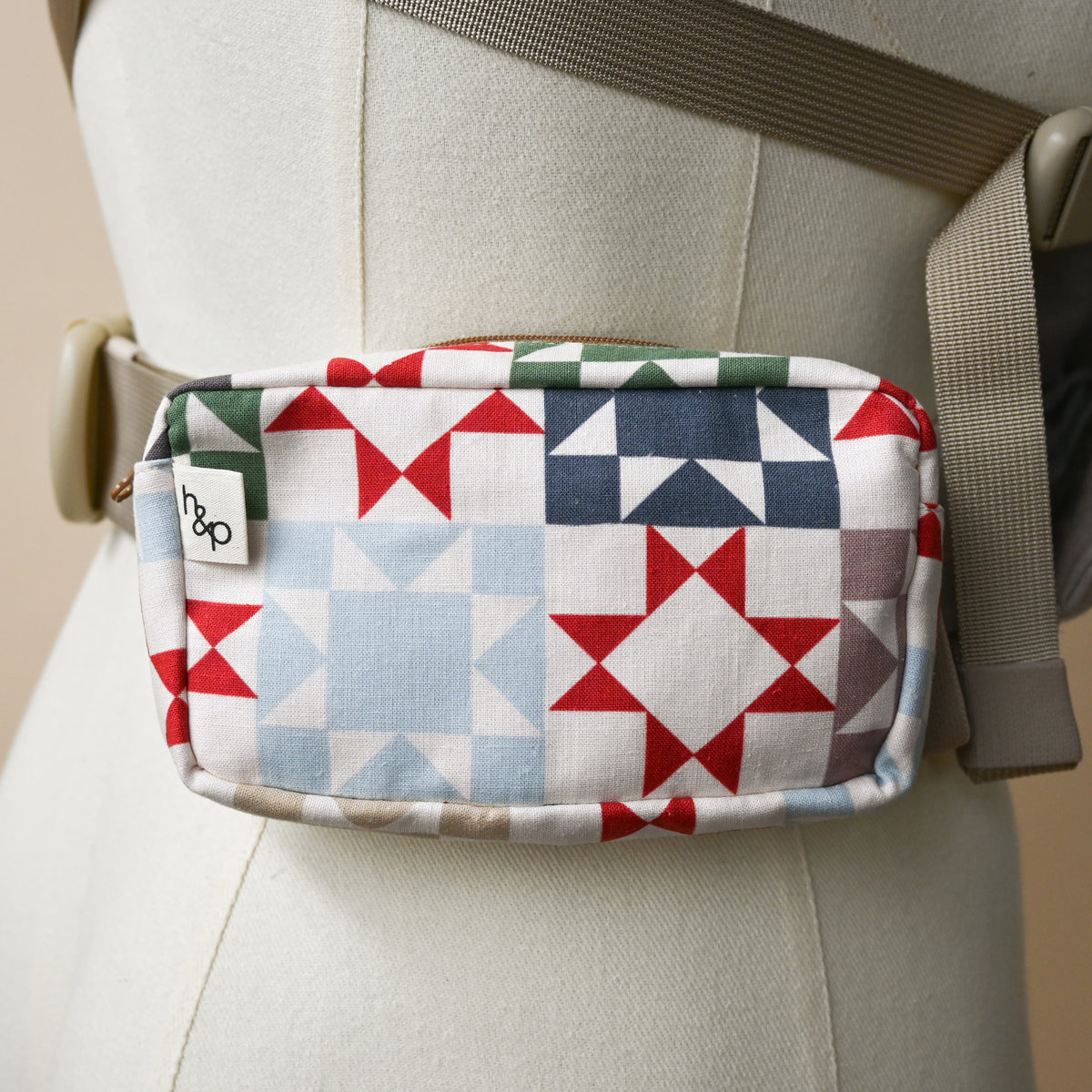 Carrier Pocket in Heirloom, a zip pouch on a carrier waistband shown on a dress form, cream fabric printed with patchwork quilt star blocks in red, green, navy, slate blue and taupe.