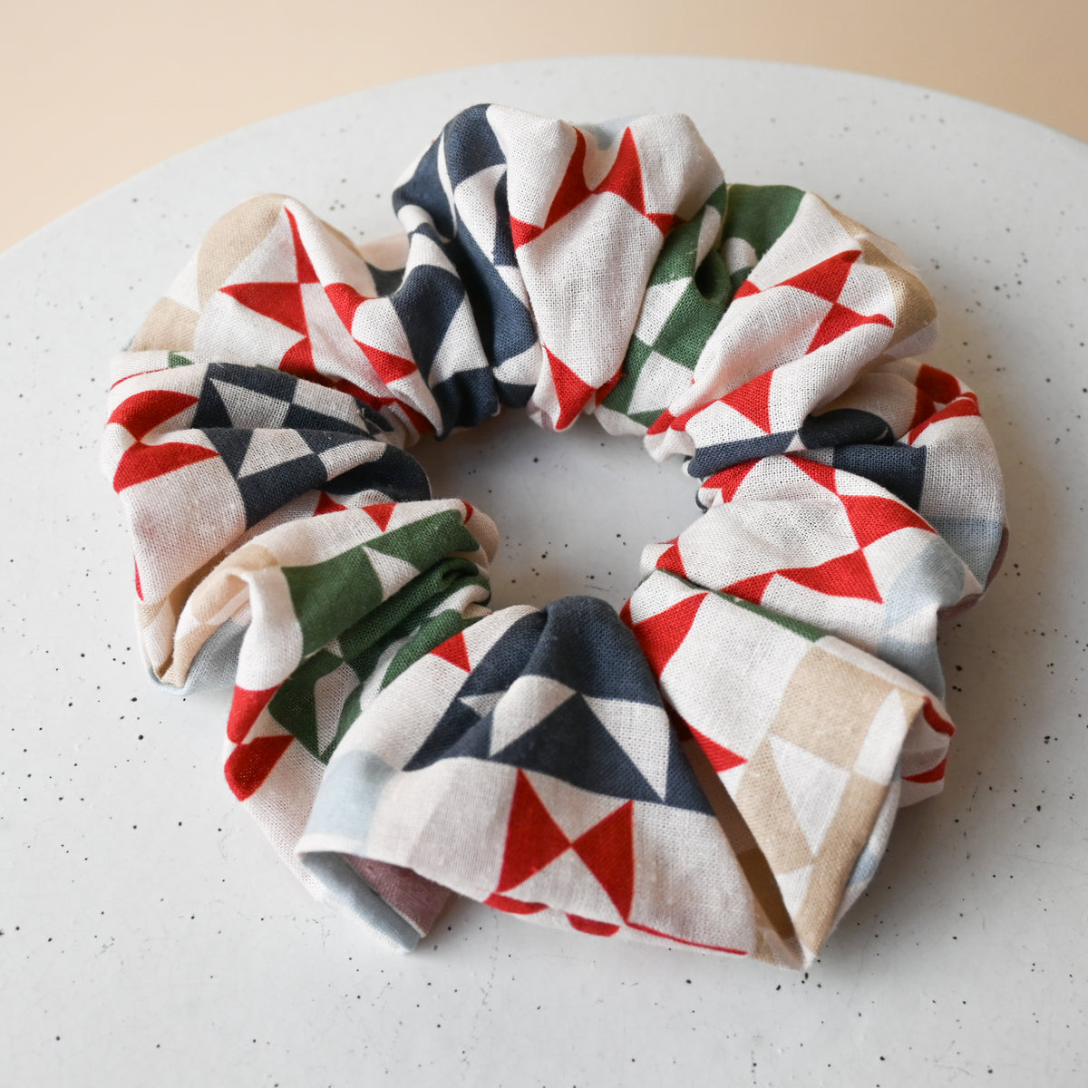 A single oversized scrunchie in Heirloom, gathered into a full, voluminous ring, cream fabric printed with patchwork quilt star blocks in red, green, navy, slate blue and taupe.