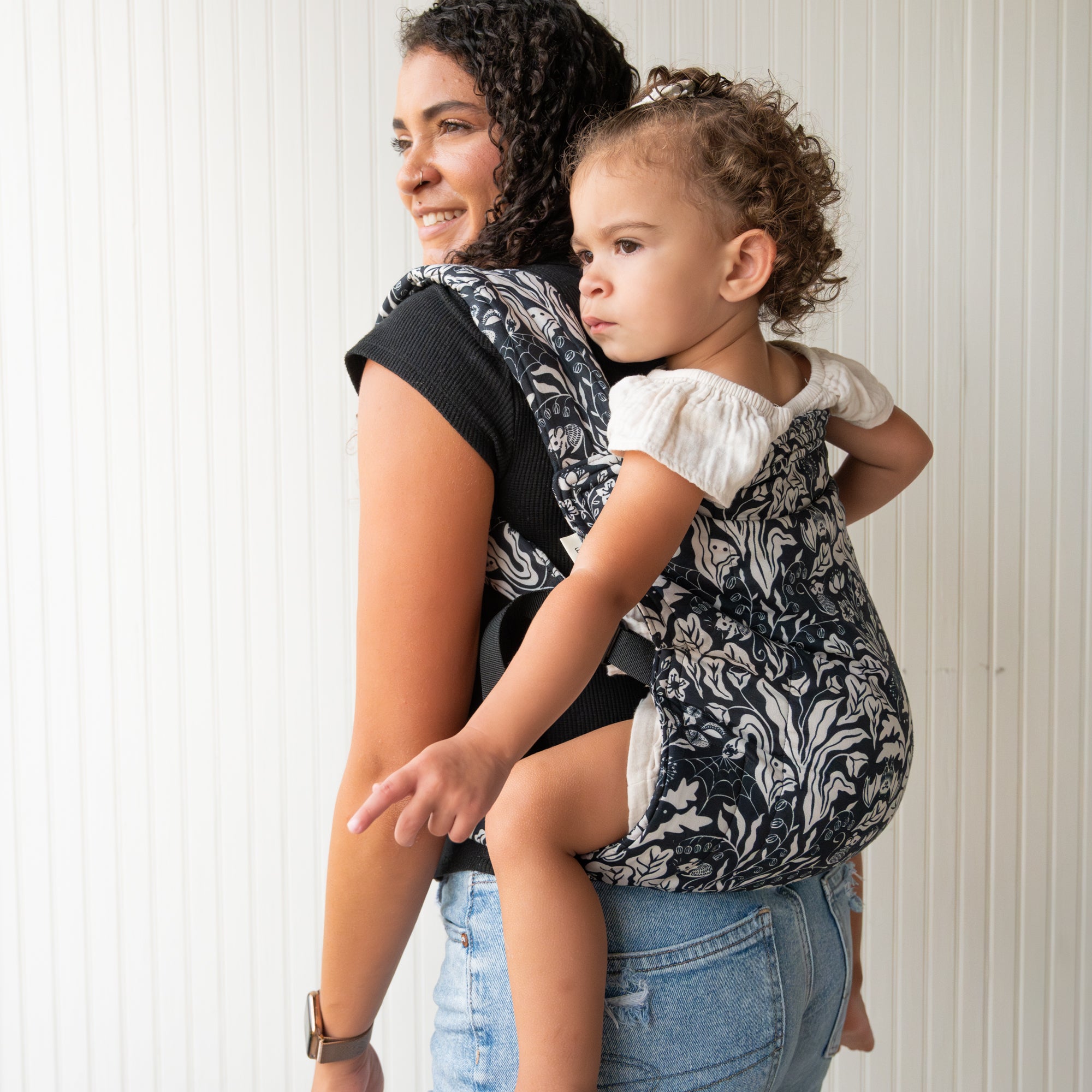 Woman carrying a child in a black and white floral baby carrier against a white wall.