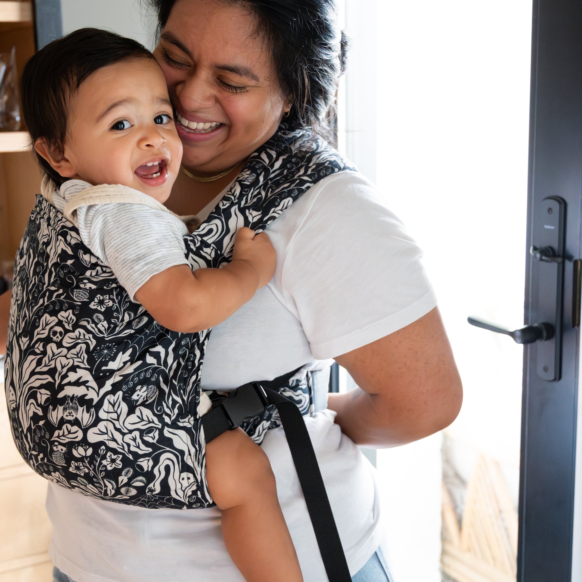Woman holding a baby in a carrier, both smiling indoors.