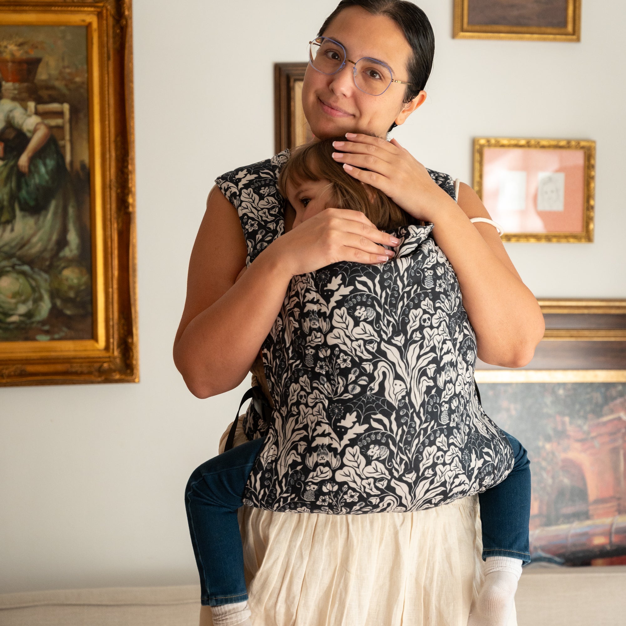 Woman carrying a child on her back with a floral witchy patterned carrier, indoors.