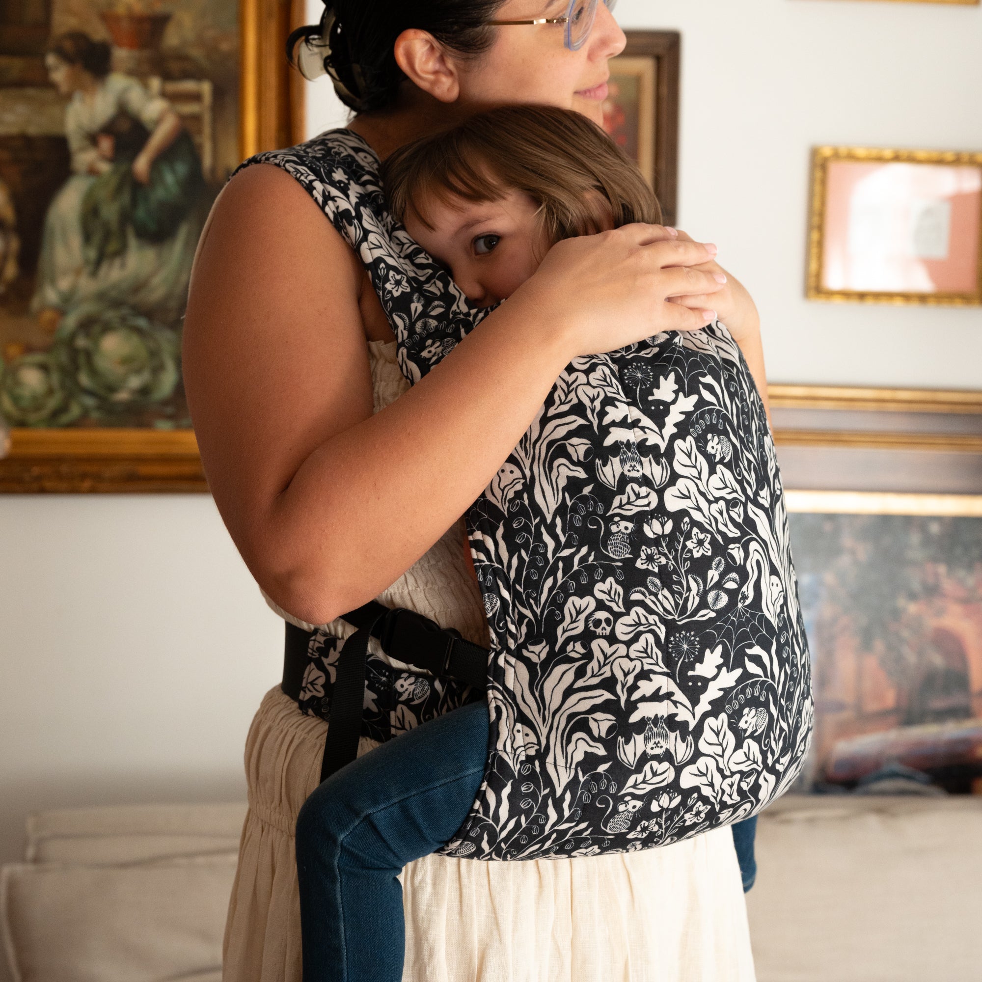 Woman carrying a child on her back with a floral witchy patterned carrier, indoors.