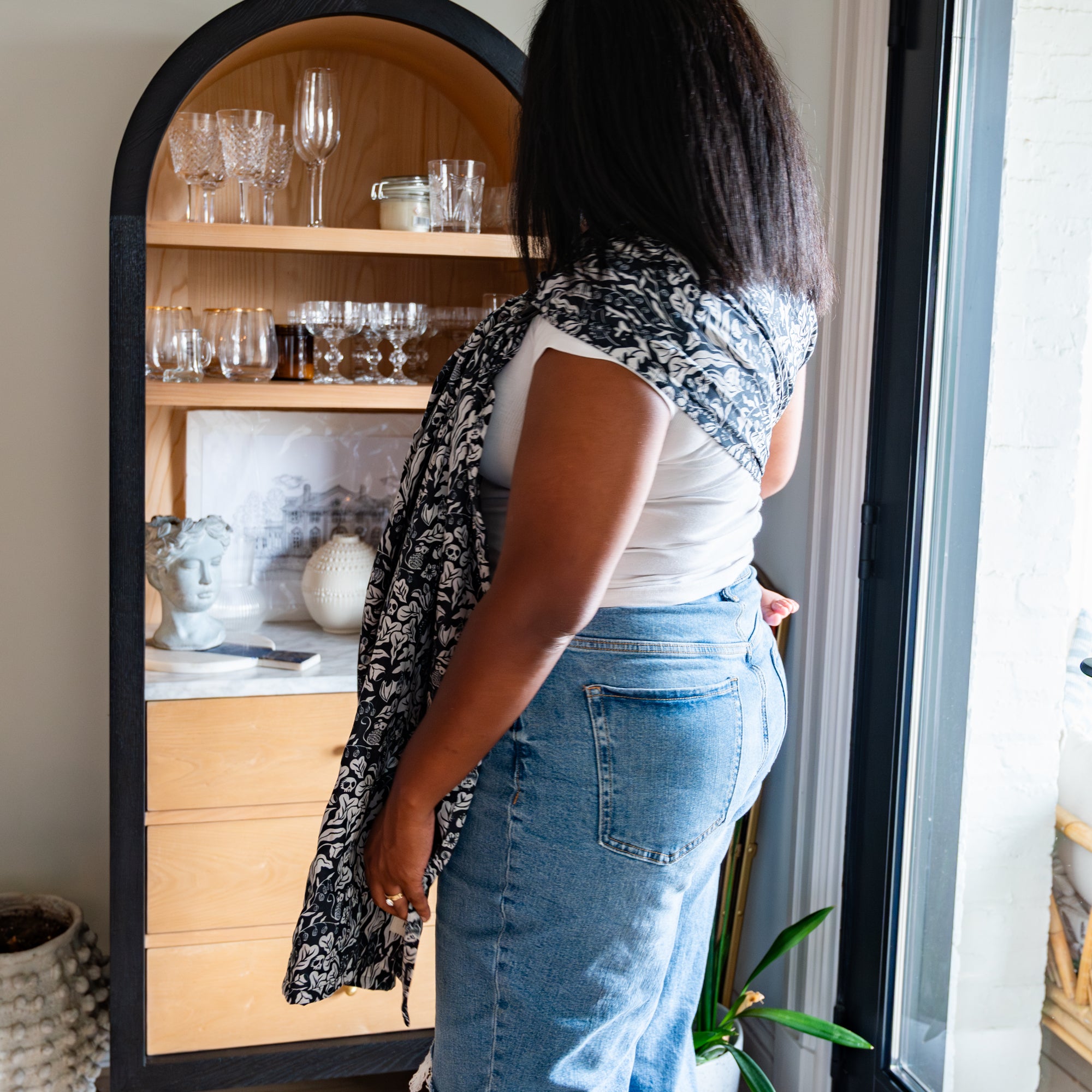 Person wearing a white top and blue jeans standing in front of a wooden cabinet with glassware while wearing a baby in a witchy pattern ring sling carrier. 