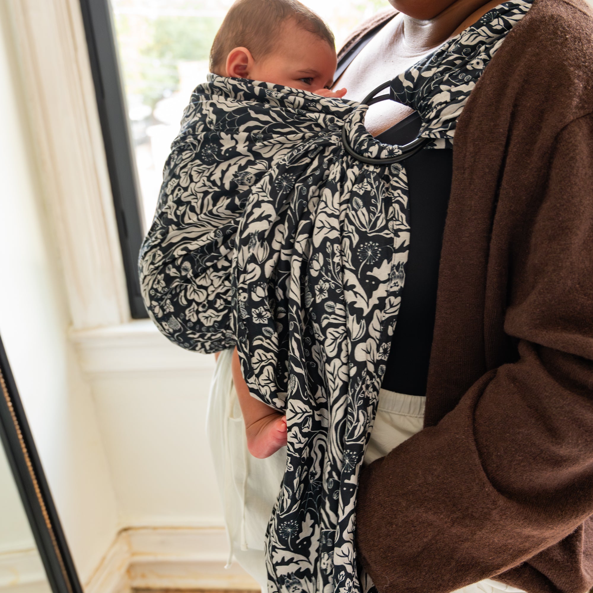 Person holding a baby wrapped in a patterned cloth ring sling baby carrier indoors.