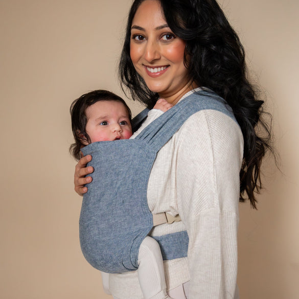 Woman holding a baby in a blue newborn soft carrier against a beige background
