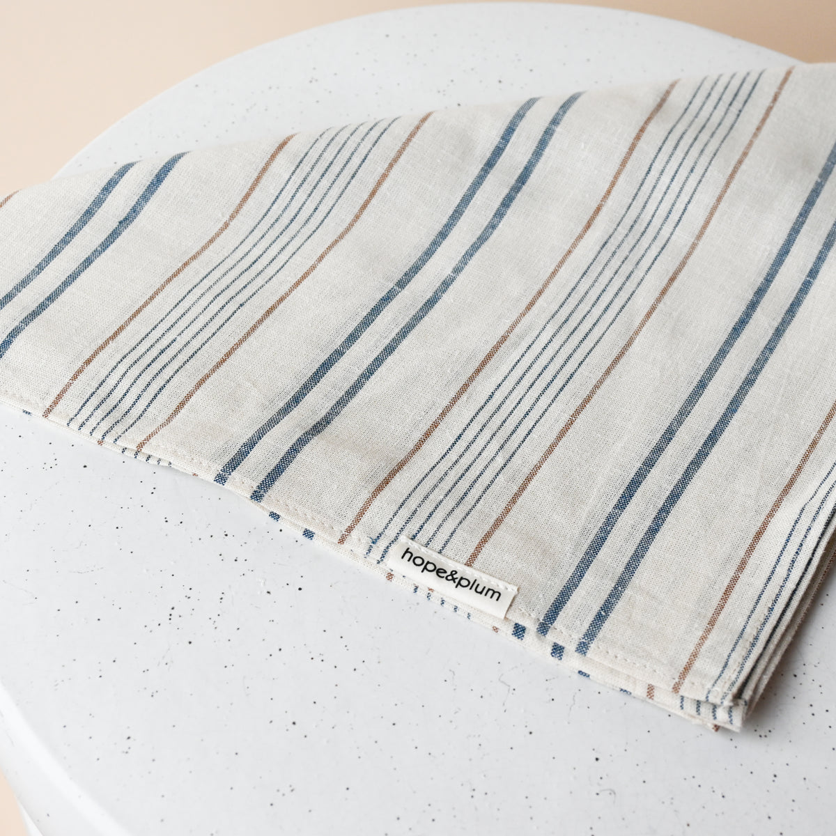 Large Bandana in Lugano, folded on a white table, cream woven chambray with blue stripes and fine rust pinstripes.