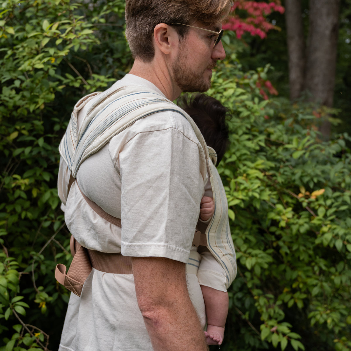 A wearer carries a baby in the Lugano Lark Baby Carrier outdoors, seen from the side, the woven navy and tan striped chambray across the shoulder.