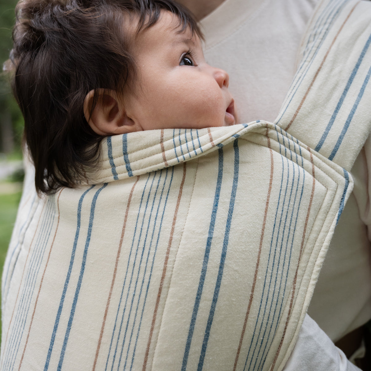 A baby looks out over the folded headrest of the Lugano Lark Baby Carrier, showing the woven stripes of navy and caramel on cream chambray.