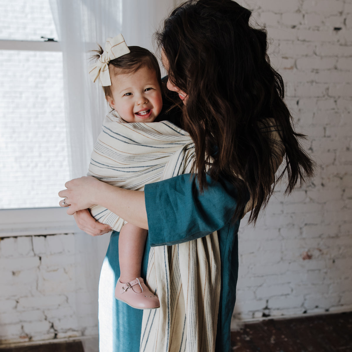 Side view of a smiling baby seated in the Lugano Ring Sling, the woven navy and tan striped chambray supporting them against the wearer's chest.
