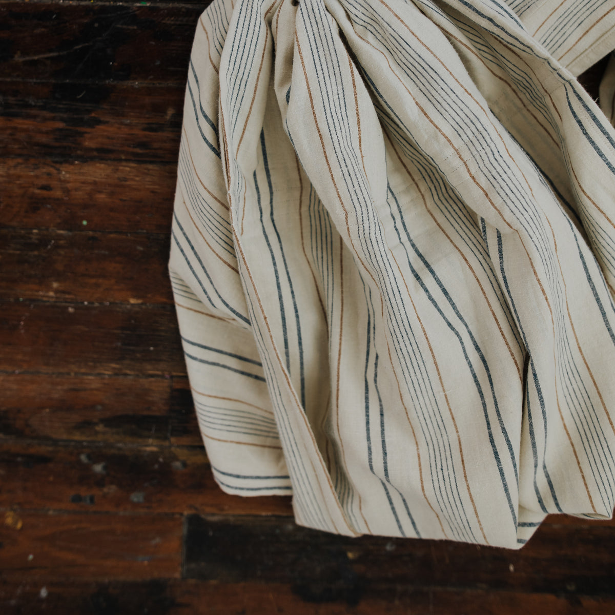 The Lugano chambray laid out on a wooden floor, its woven stripes of slate navy and caramel tan running the length of the cream fabric.