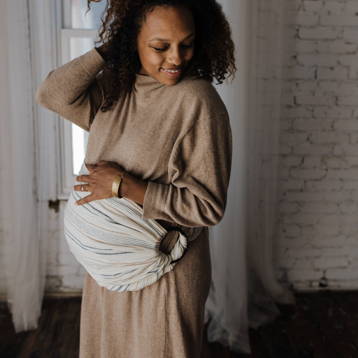 A wearer looks down at the pouch of the Lugano Ring Sling, the woven chambray falling in soft folds of navy and tan stripes on cream.