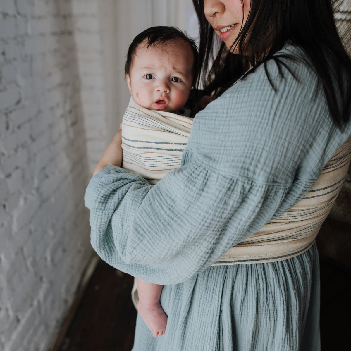 A baby looks out from the Lugano Ring Sling held against the wearer's chest, the woven navy and tan stripes visible across the sling and over the shoulder.