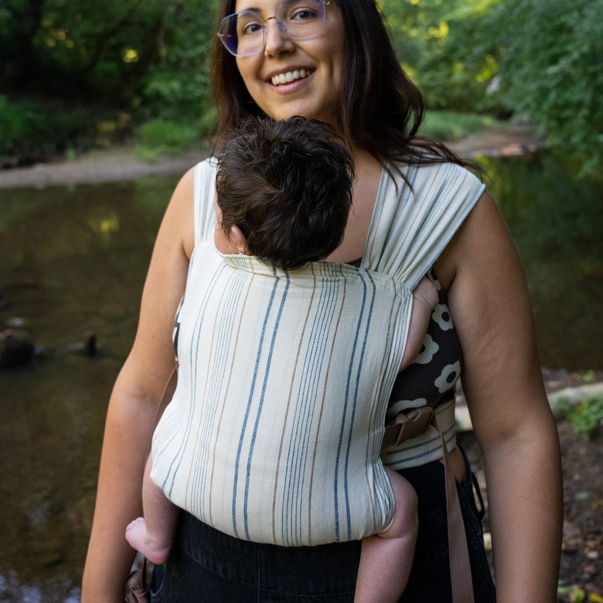 Lugano Sprout Newborn Carrier worn in a front carry beside a creek, a baby's head resting against the panel of woven navy and tan stripes on cream chambray.