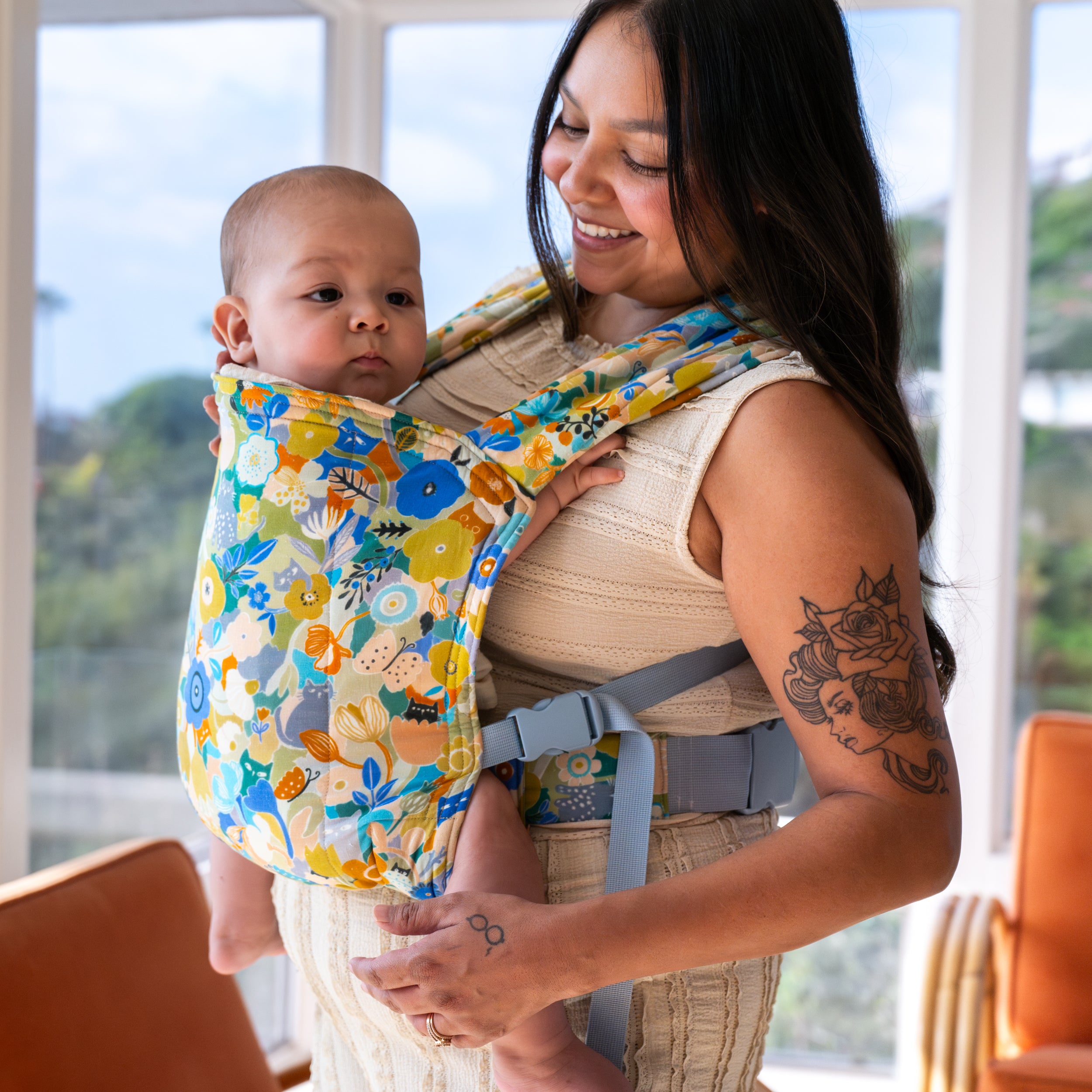 Woman holding a baby in a colorful buckle carrier indoors.