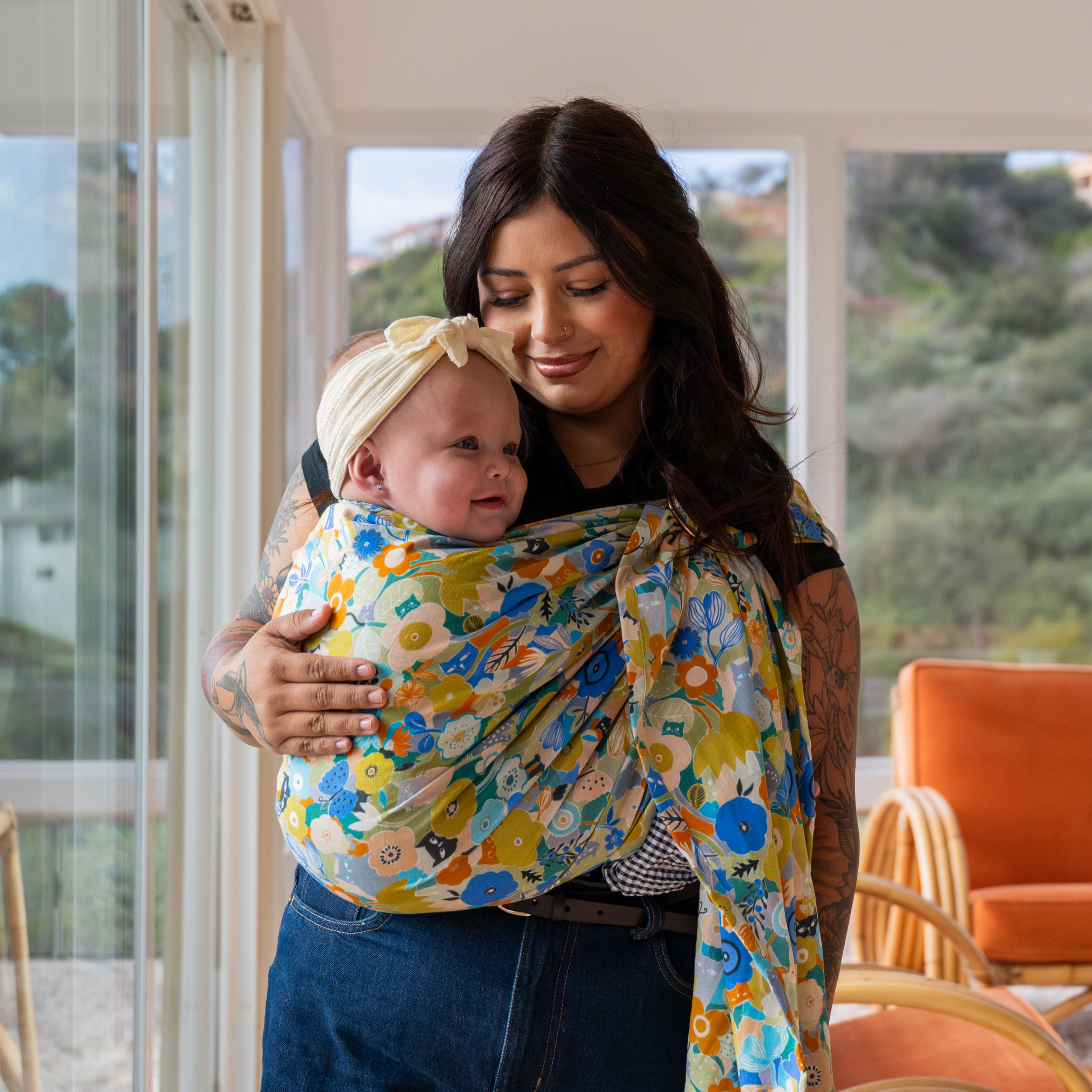 Woman holding a baby wrapped in a colorful floral ring sling in a home setting