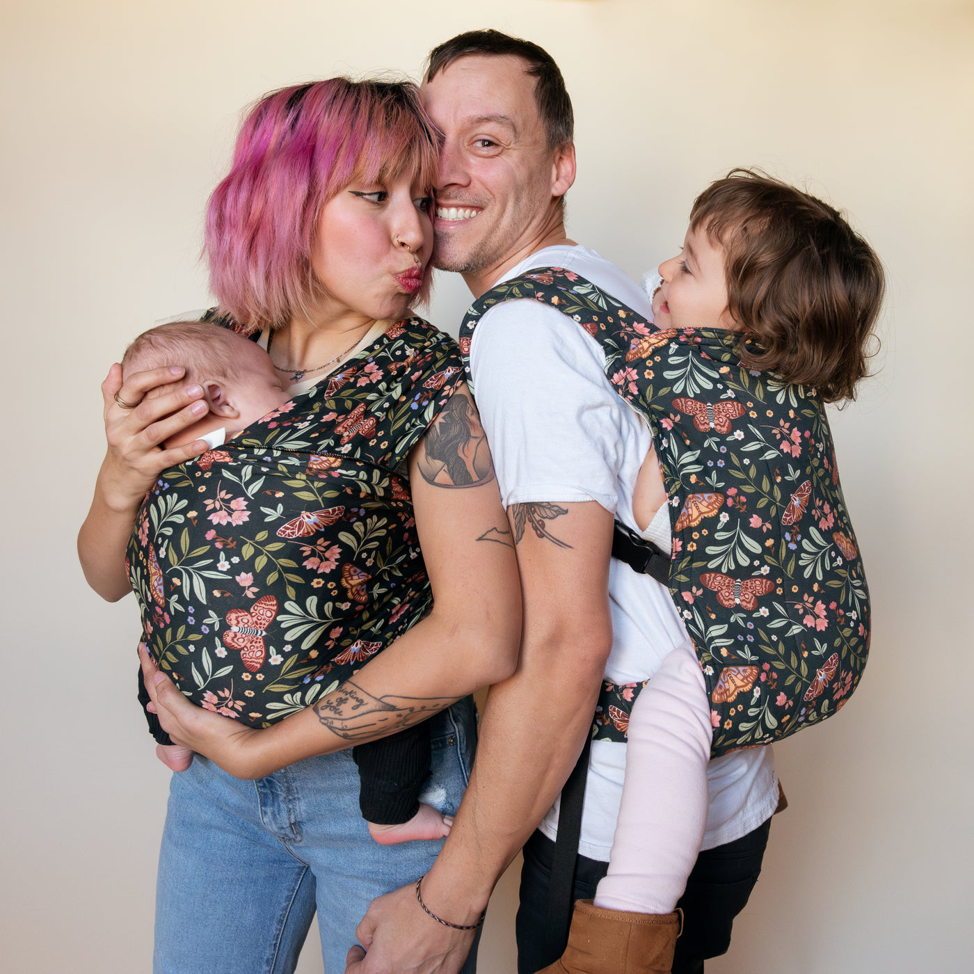 Two caregivers stand back-to-back, each carrying a child in matching Night Garden carriers — a dark botanical print with moths, pink flowers, and green leaves — one in a baby wrap with a newborn, one in a Lark with a toddler.