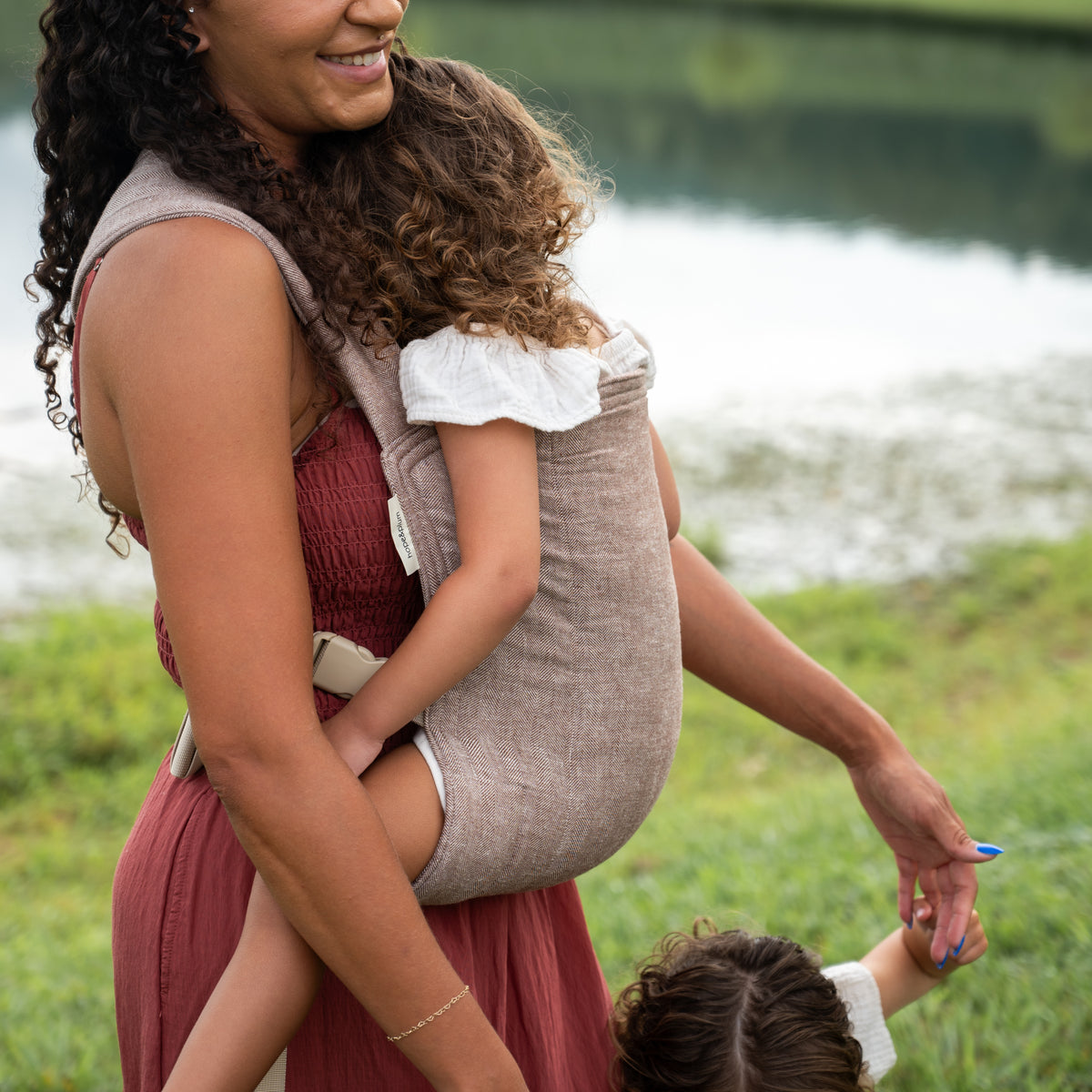 Smiling mother carrying a toddler on her back in the Oak Lark Baby Carrier, a chocolate and oatmeal herringbone weave, beside a pond.