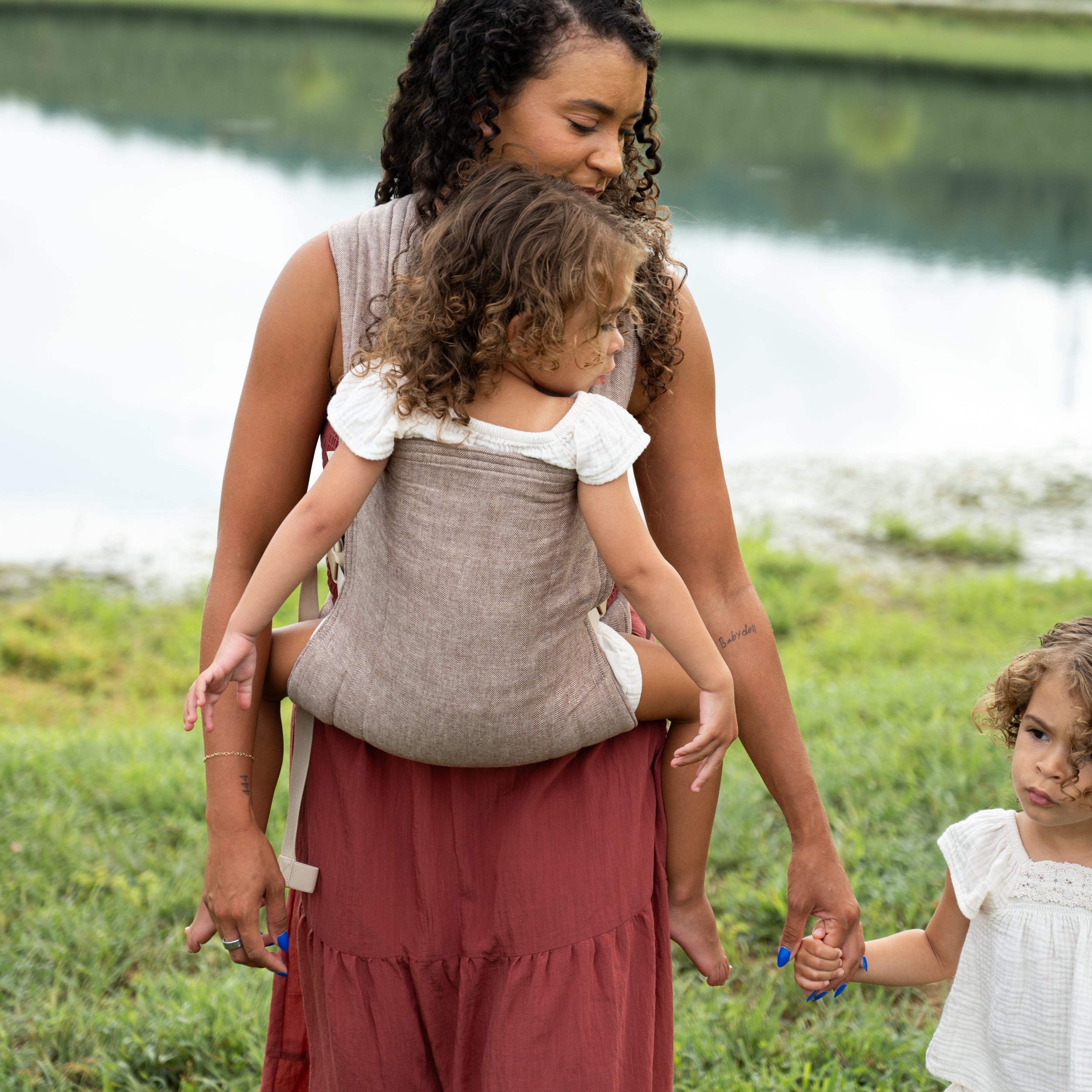 Mother wearing the Oak Lark Baby Carrier, a chocolate and oatmeal herringbone weave, on her back with a toddler inside, walking hand-in-hand with another child near a lake.