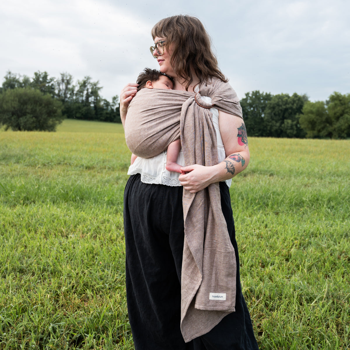 Woman standing in a field cradling a sleeping newborn against her chest in the Oak Ring Sling, a chocolate and oatmeal herringbone weave, with the fabric gathered through the ring at her shoulder.