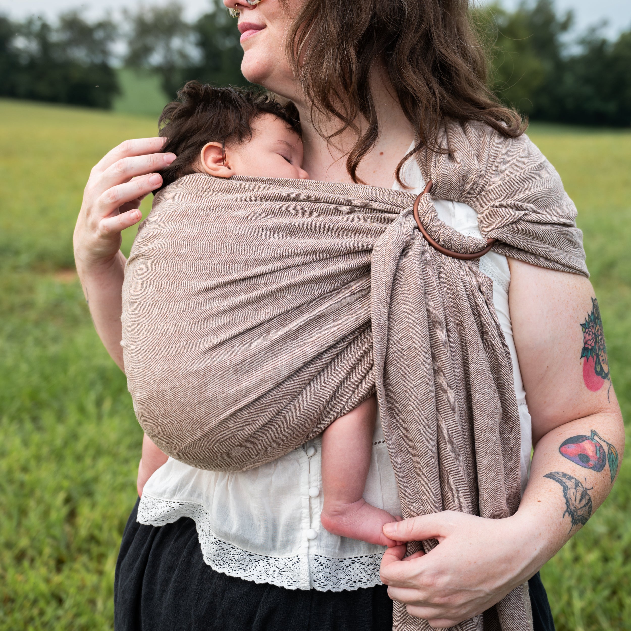 Close-up of a woman holding a sleeping newborn wrapped in the Oak Ring Sling's chocolate and oatmeal herringbone weave, showing the shoulder ring.
