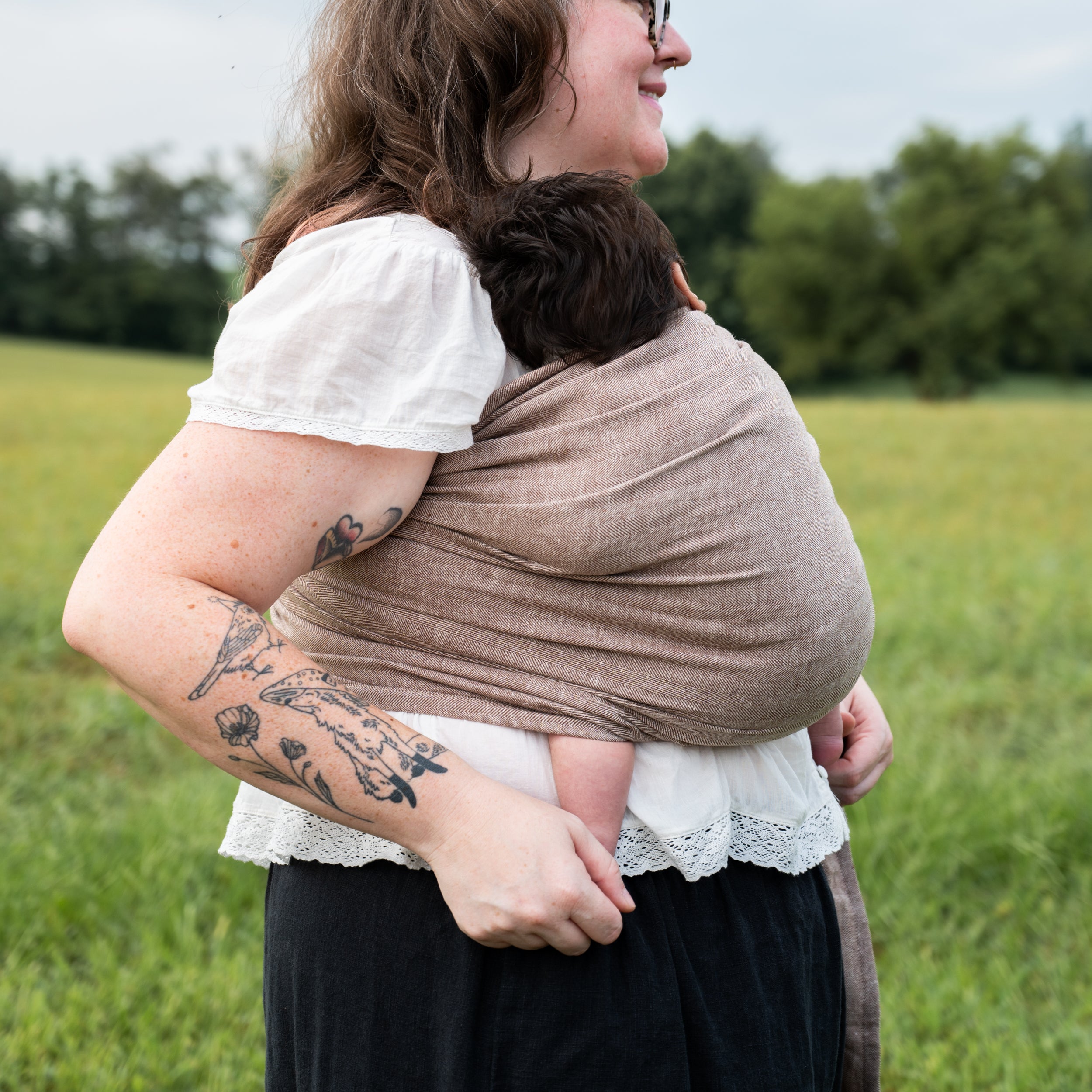 Smiling woman carrying a baby against her chest in the Oak Ring Sling, a chocolate and oatmeal herringbone weave, standing in a grassy field.
