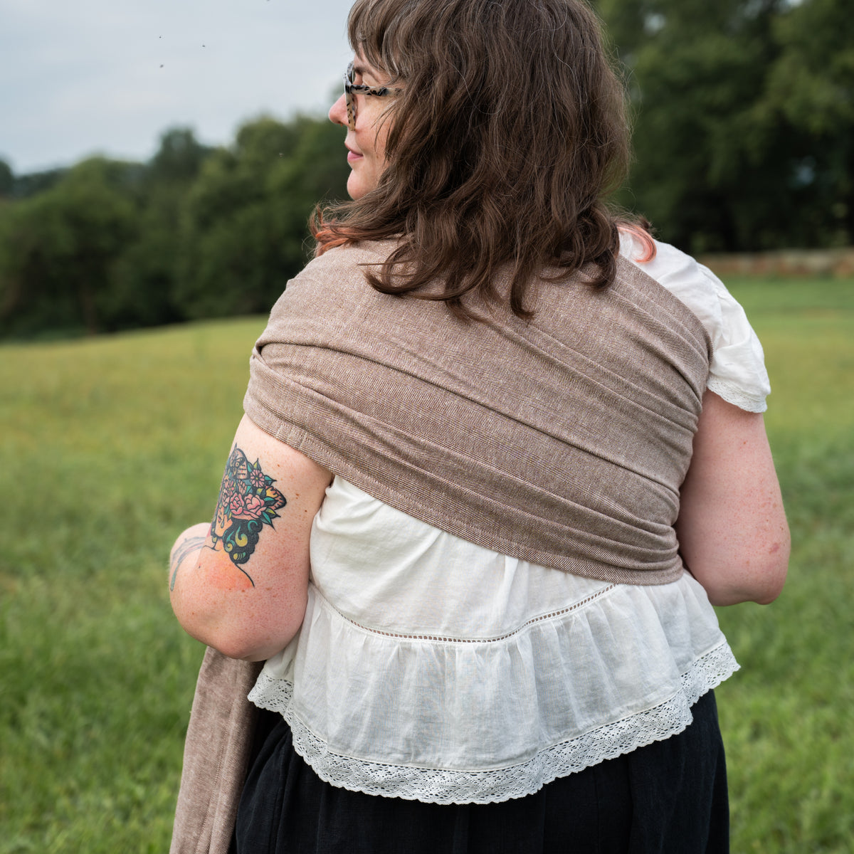 Back view of a woman wearing the Oak Ring Sling, a chocolate and oatmeal herringbone weave, over one shoulder, smiling, standing in a field.