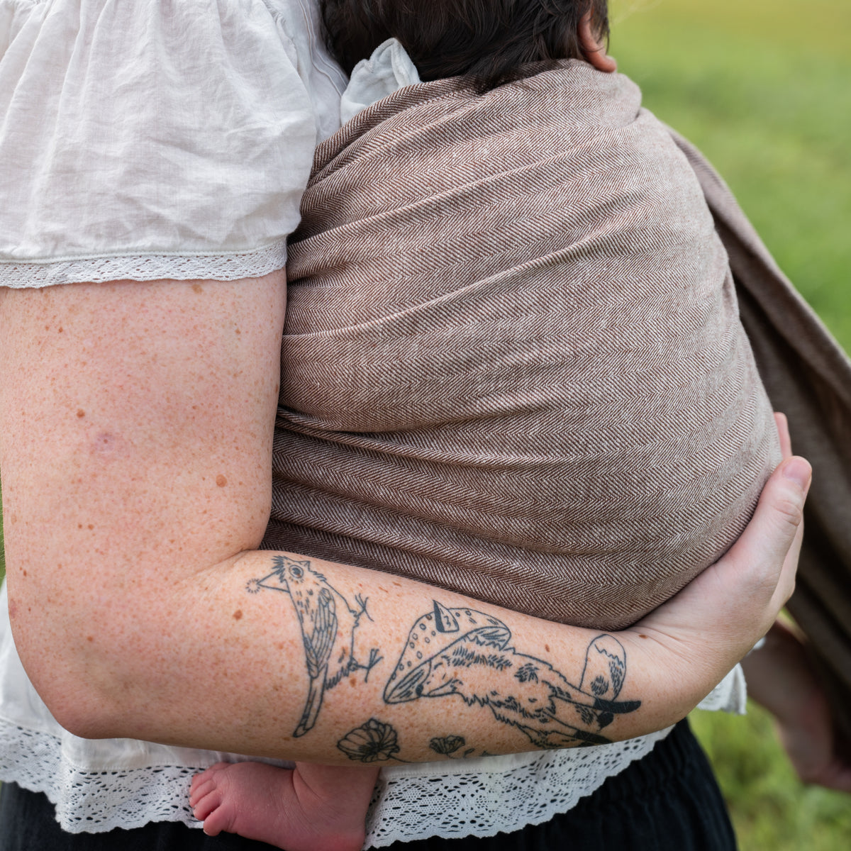 Close-up of the Oak Ring Sling's chocolate and oatmeal herringbone weave wrapped around a woman's back while she holds a baby.
