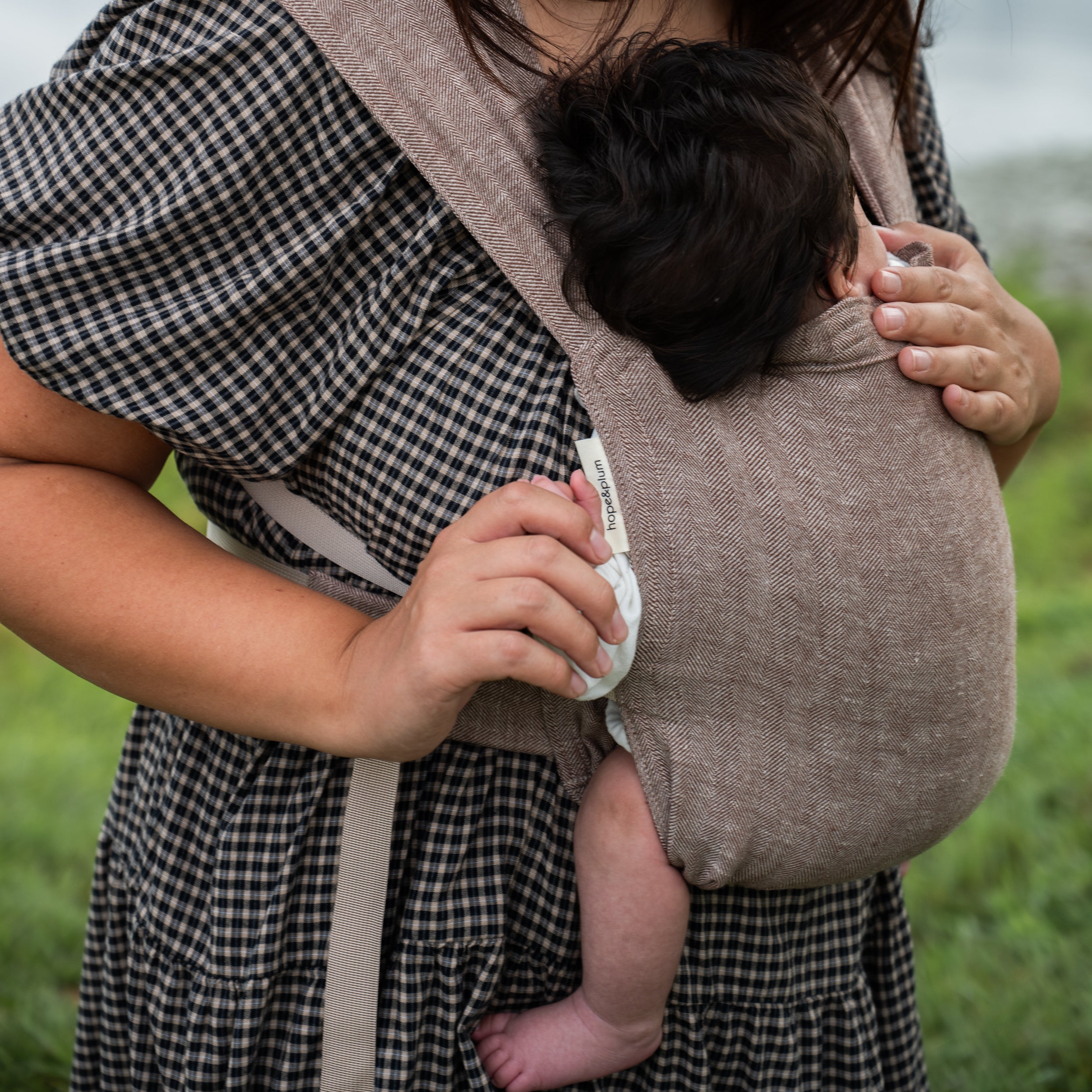 Close-up of a newborn's head resting against a woman's chest in the Oak Sprout Newborn Carrier's chocolate and oatmeal herringbone weave, her hand supporting the baby's back.