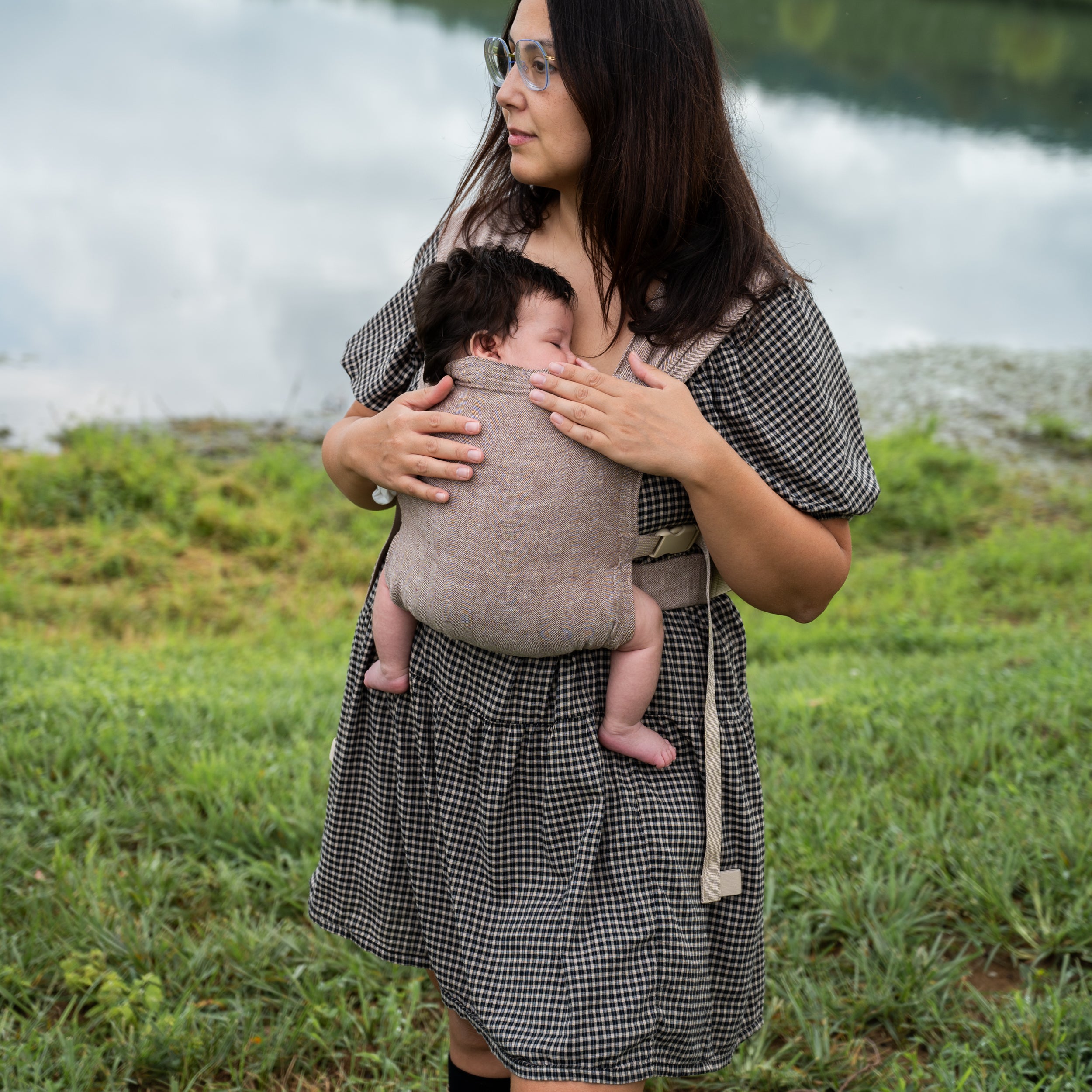 Woman with glasses looking down at a sleeping newborn worn on her chest in the Oak Sprout Newborn Carrier, a chocolate and oatmeal herringbone weave, standing by a lake.