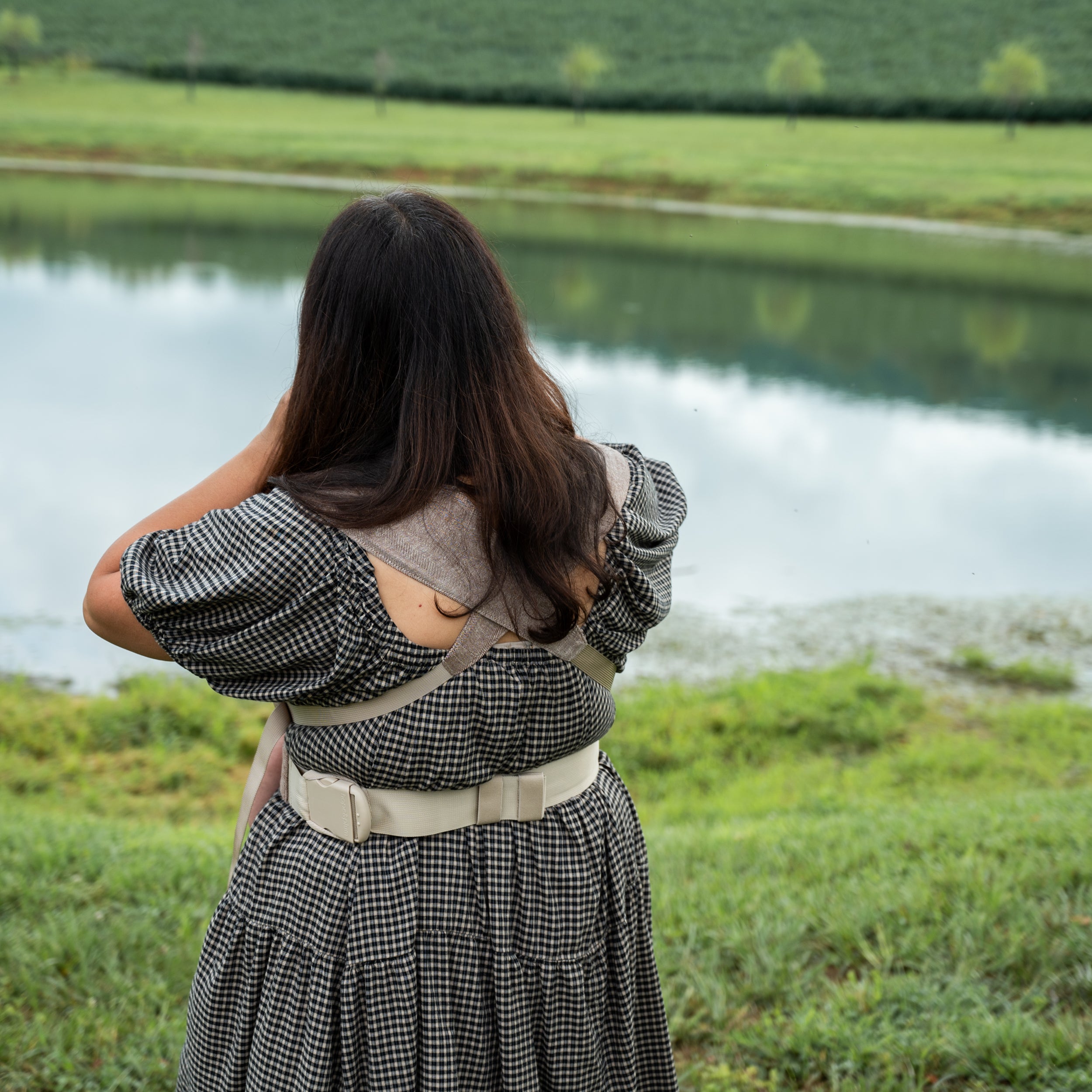 Back view of a woman wearing the Oak Sprout Newborn Carrier, a chocolate and oatmeal herringbone weave, one hand resting on her shoulder, standing by a lake.