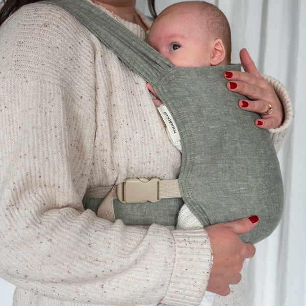 Sprout Newborn Carrier in Palm — close-up of a newborn peeking out of the heathered sage-olive chambray carrier, caregiver's hand resting on the panel.