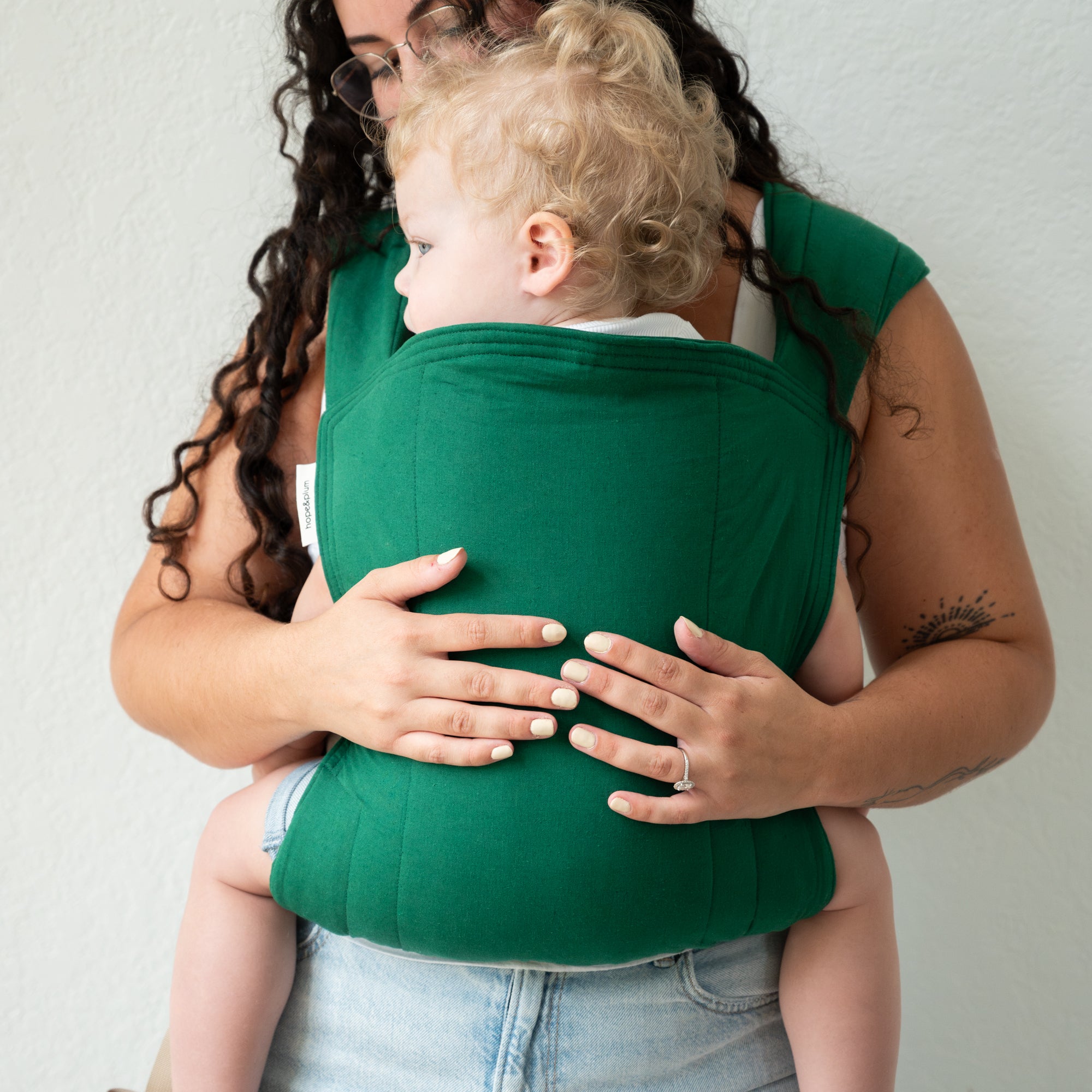 Woman holding a child in a green baby carrier against a plain background