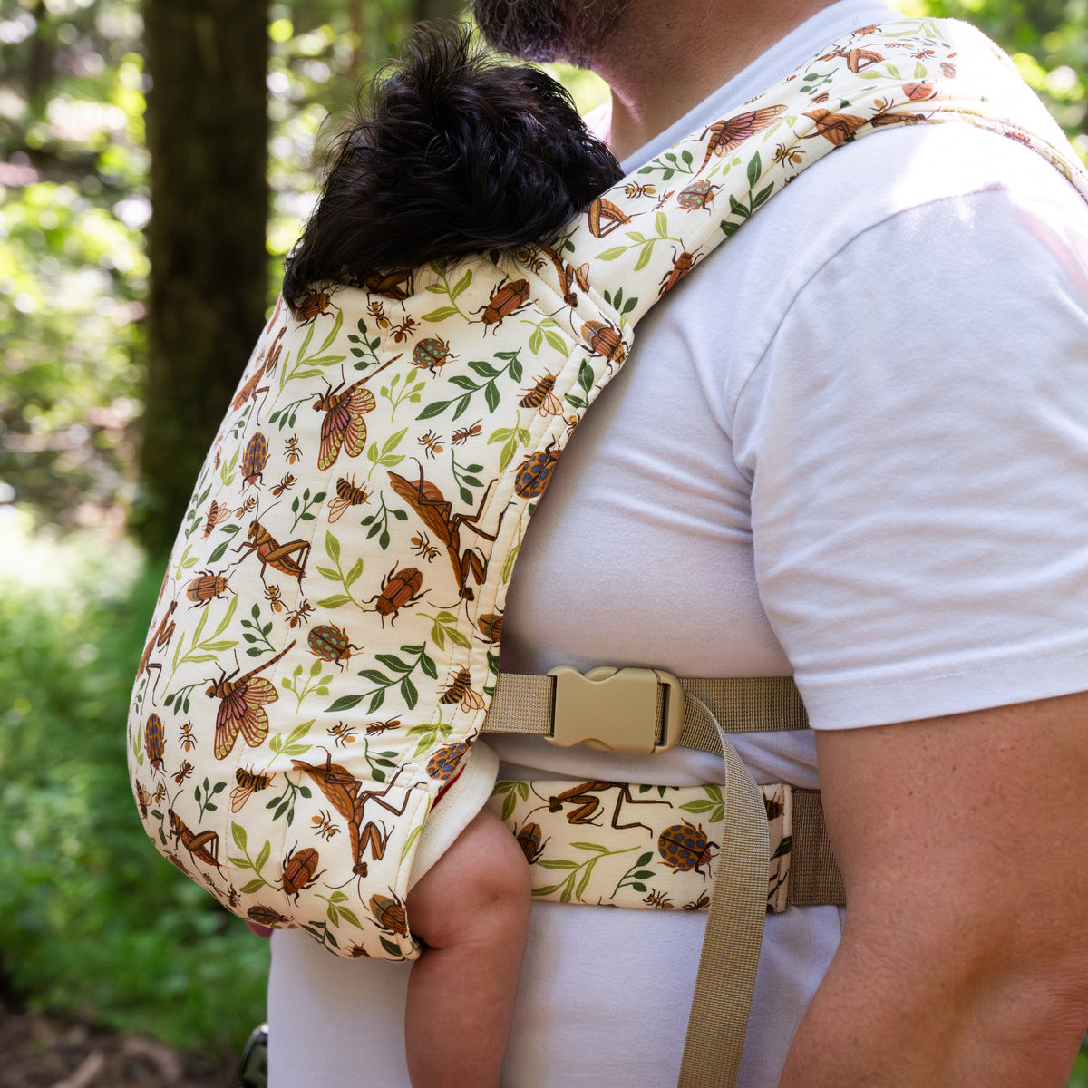 Close-up of a sleeping toddler worn on a man's chest in the Reed Lark Baby Carrier, a natural hemp-cotton fabric printed with warm brown bugs and leaves, showing ladybug, beetle, and dragonfly details.