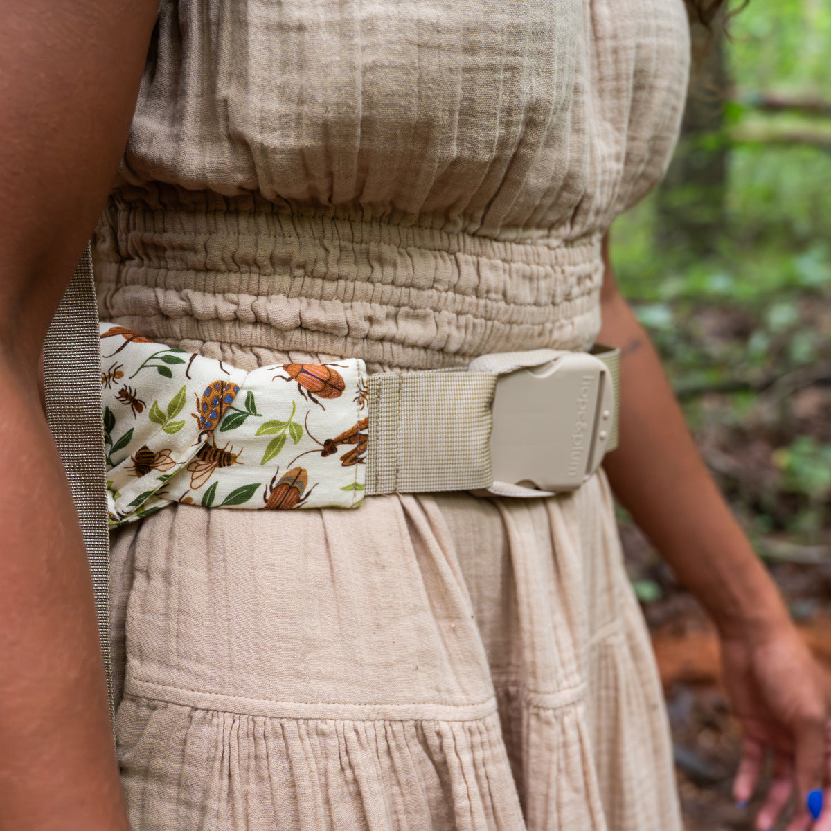 Close-up of the Reed Lark Kid Carrier's waist buckle and hope&plum label, showing the natural hemp-cotton fabric printed with warm brown bugs and leaves.