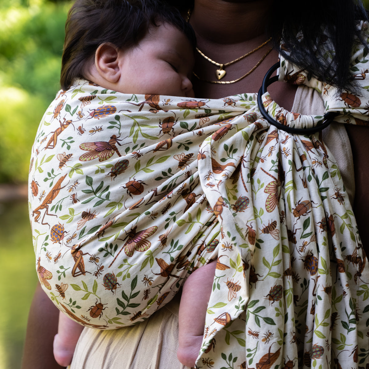 Close-up of a baby cradled against a woman's chest in the Reed Ring Sling, a natural hemp-cotton fabric printed with warm brown bugs and leaves, with the shoulder ring visible.