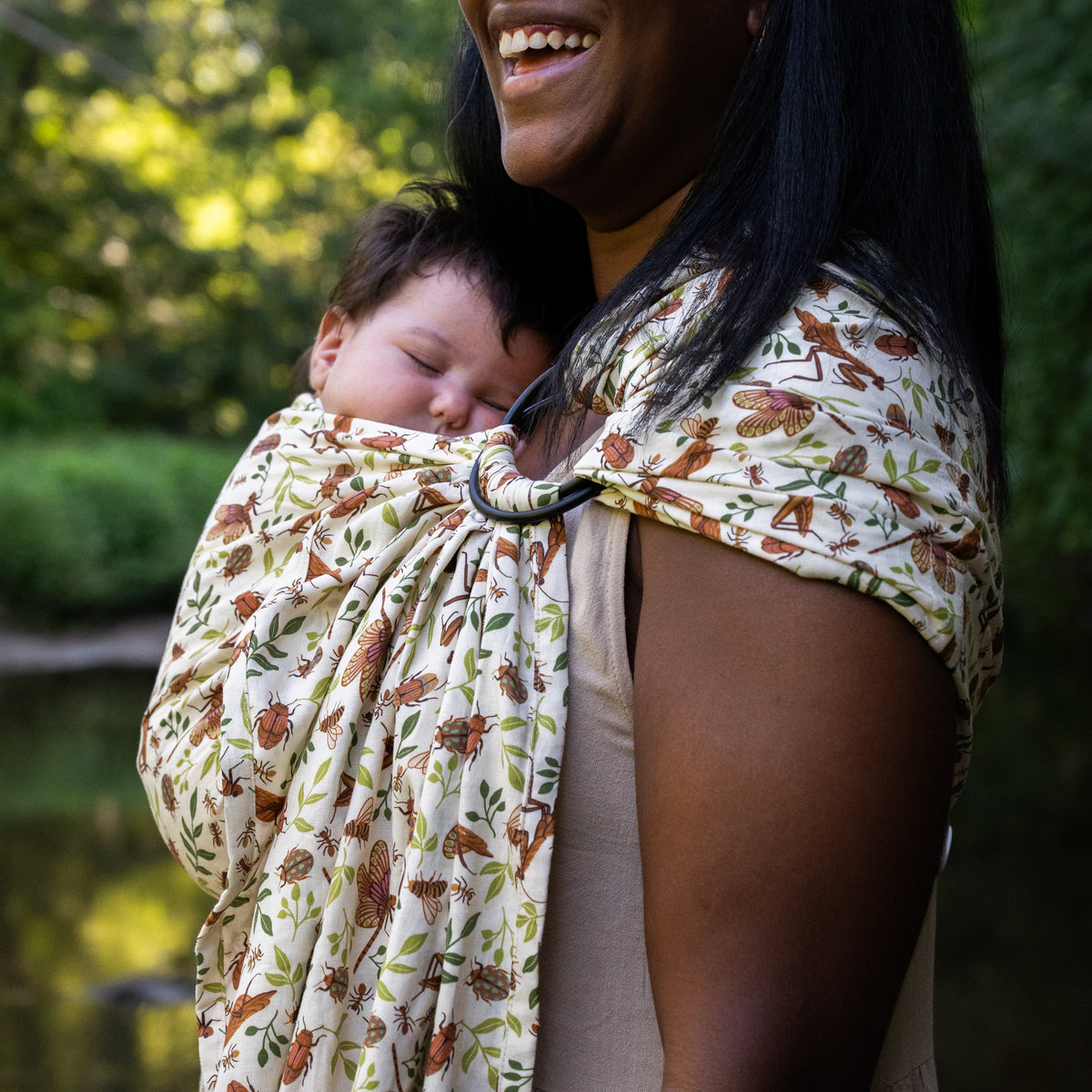 Smiling woman carrying a sleeping baby against her chest in the Reed Ring Sling, a natural hemp-cotton fabric printed with warm brown bugs and leaves.