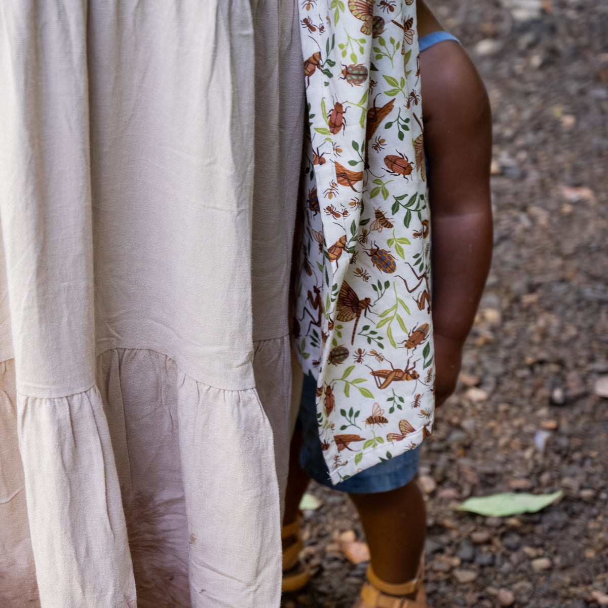Close-up of the Reed Ring Sling's tail fabric, a natural hemp-cotton fabric printed with warm brown bugs and leaves, draped beside two children outdoors.