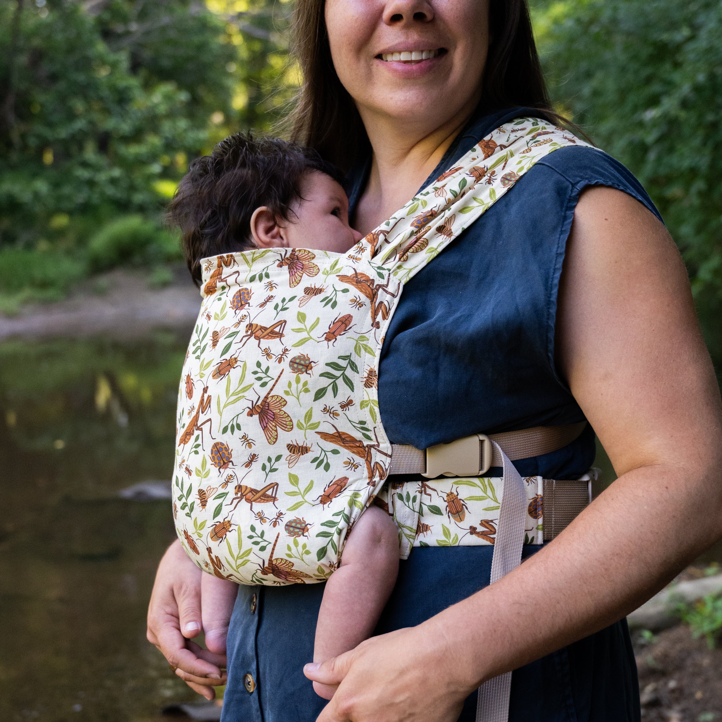 Smiling woman feeding a baby worn on her chest in the Reed Sprout Newborn Carrier, a natural hemp-cotton fabric printed with warm brown bugs and leaves, standing by a wooded creek.