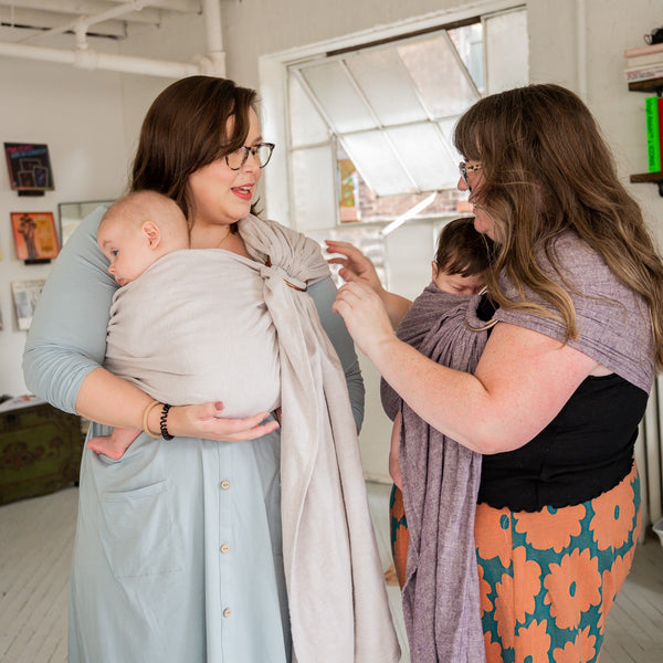 Plus-Size Mother wearing her infant in a baby carrier and being assisted by another mother babywearing in a purple baby wrap