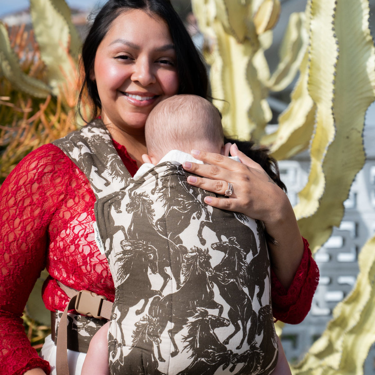 Lark Baby Carrier in Rodeo — caregiver smiling by a cactus while holding a baby in the mustang print carrier.