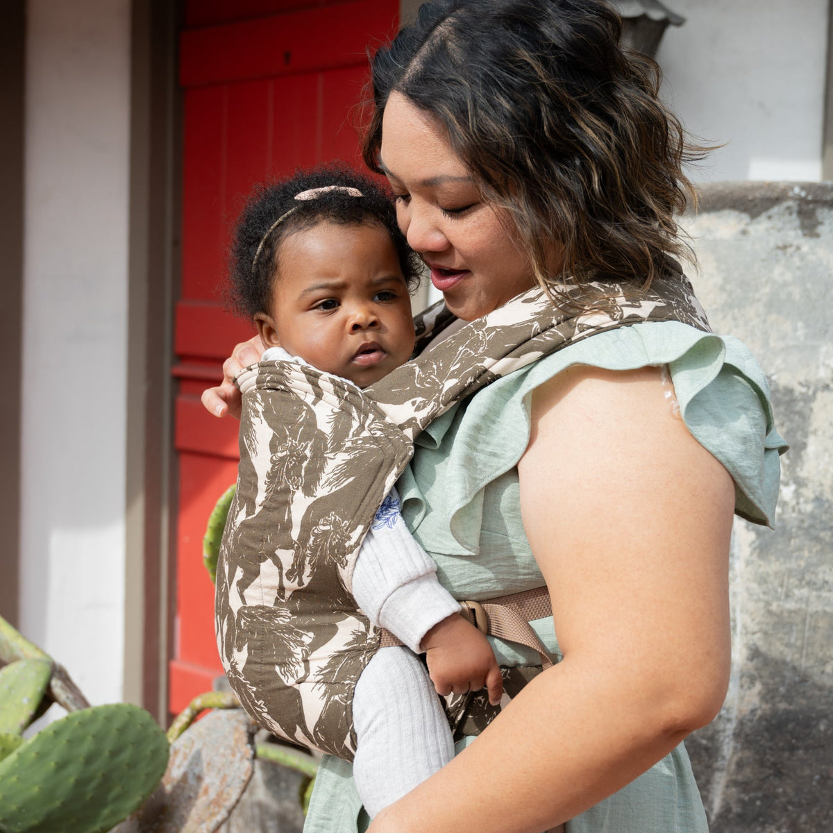 Lark Baby Carrier in Rodeo — caregiver carrying a toddler outside a house with a red door, mustang print carrier in view.