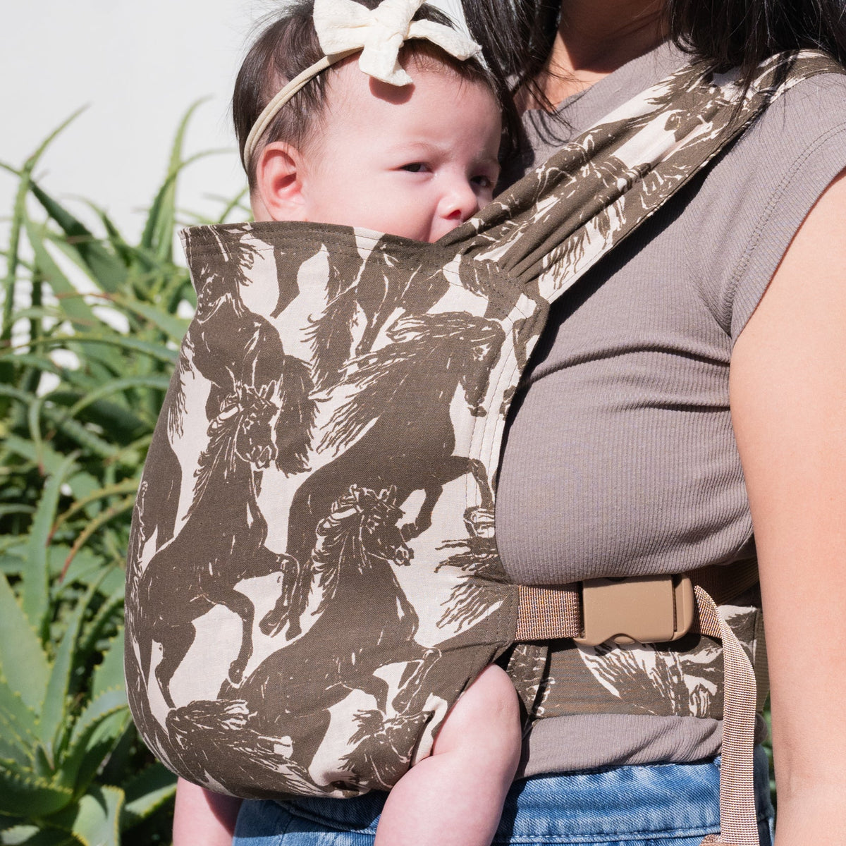 Sprout Newborn Carrier in Rodeo — caregiver looking down at a newborn in a bow headband, mustang print carrier.