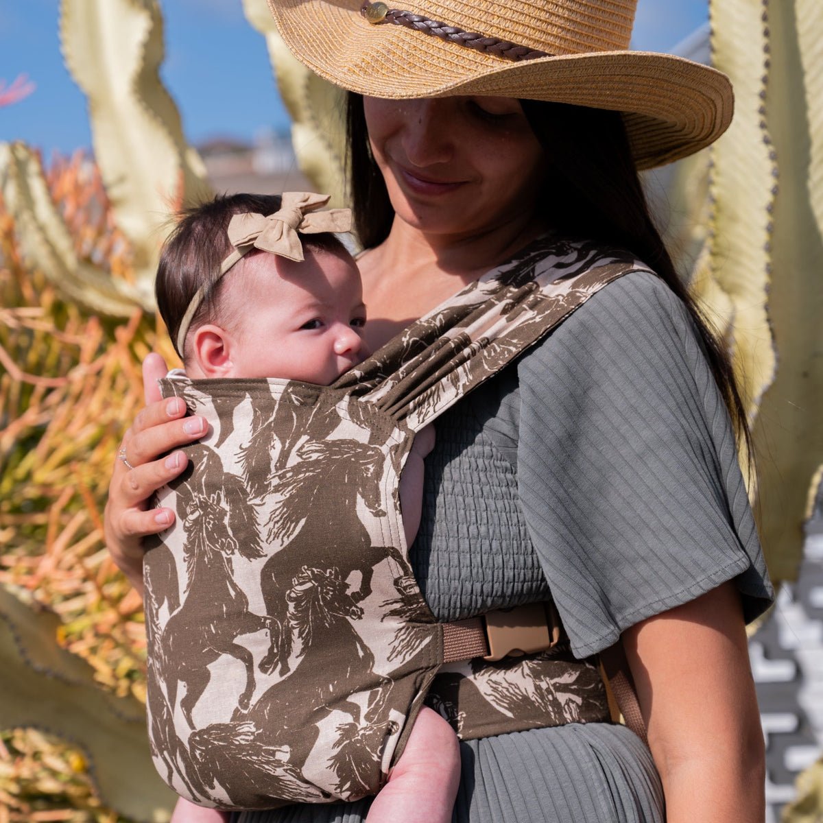 Sprout Newborn Carrier in Rodeo — caregiver in a straw hat looking aside while holding a newborn in the mustang print.