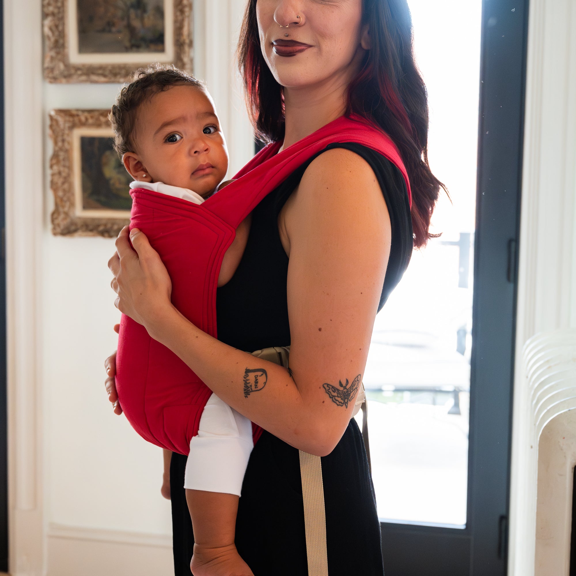 Woman holding a baby in a red baby carrier indoors.