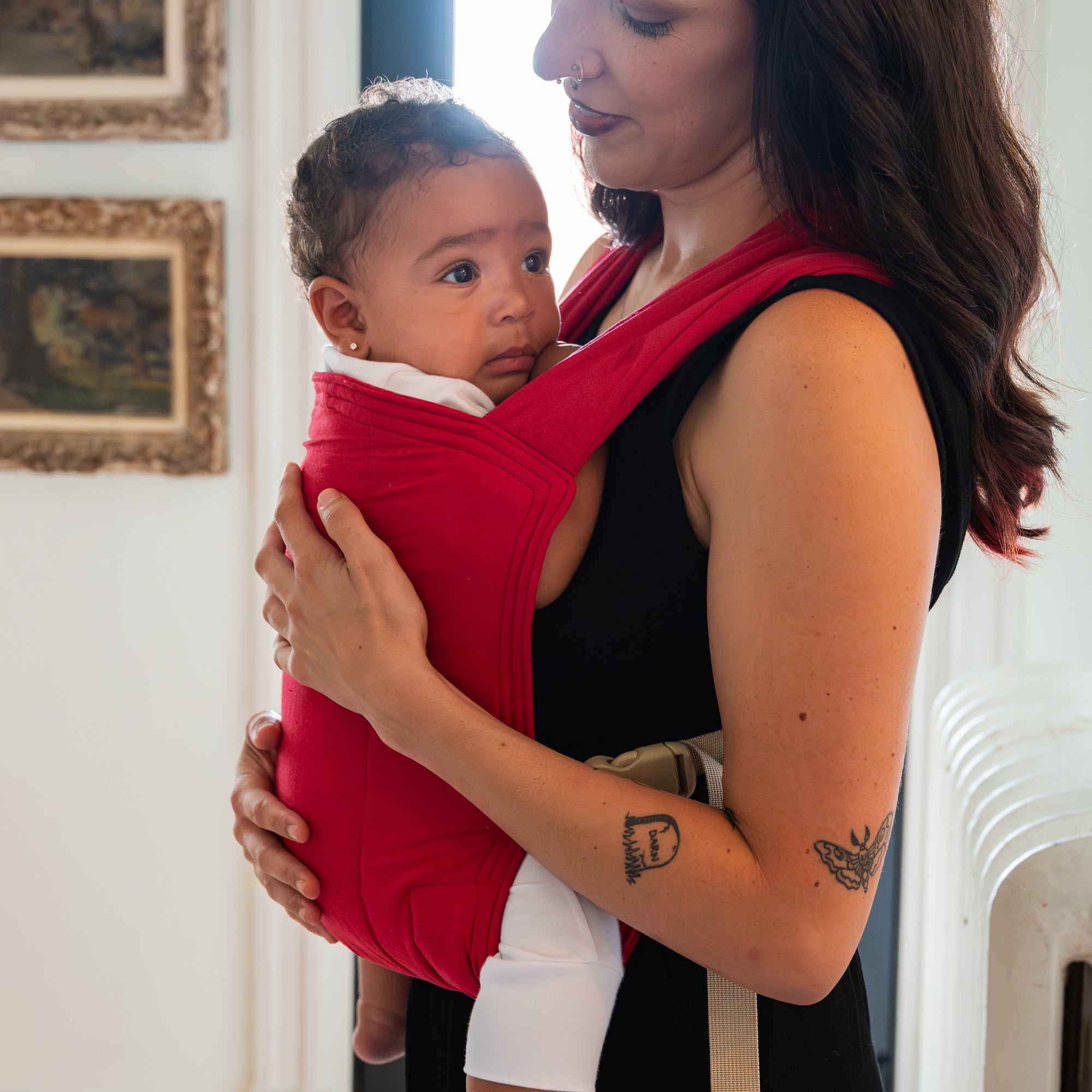 A woman holding a baby in a red soft structured baby carrier  indoors.
